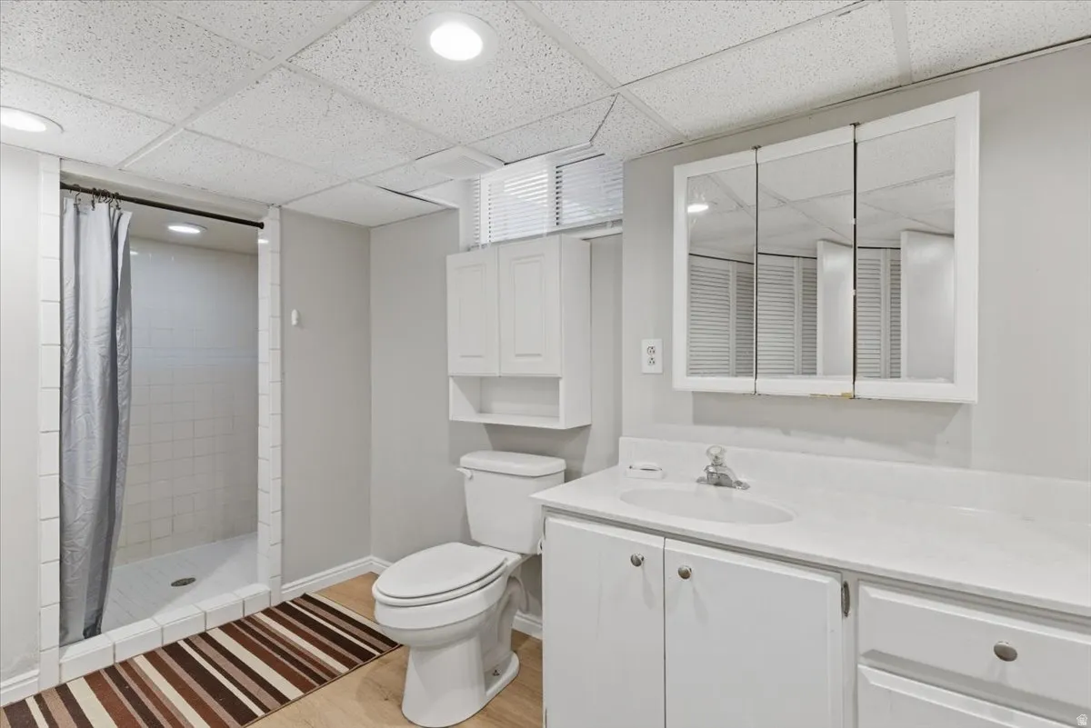 Bathroom featuring a walk-in shower with white tile surround, a white vanity with an integrated sink and white countertop, a tri-fold mirrored medicine cabinet, a toilet, and wood-finish flooring