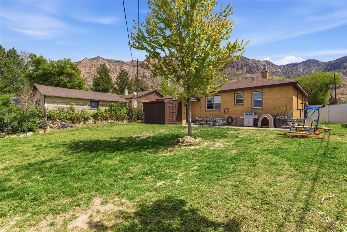 Expansive backyard featuring a manicured lawn, a detached storage shed, a brick exterior residence, mountain scenery, and a secondary long outbuilding