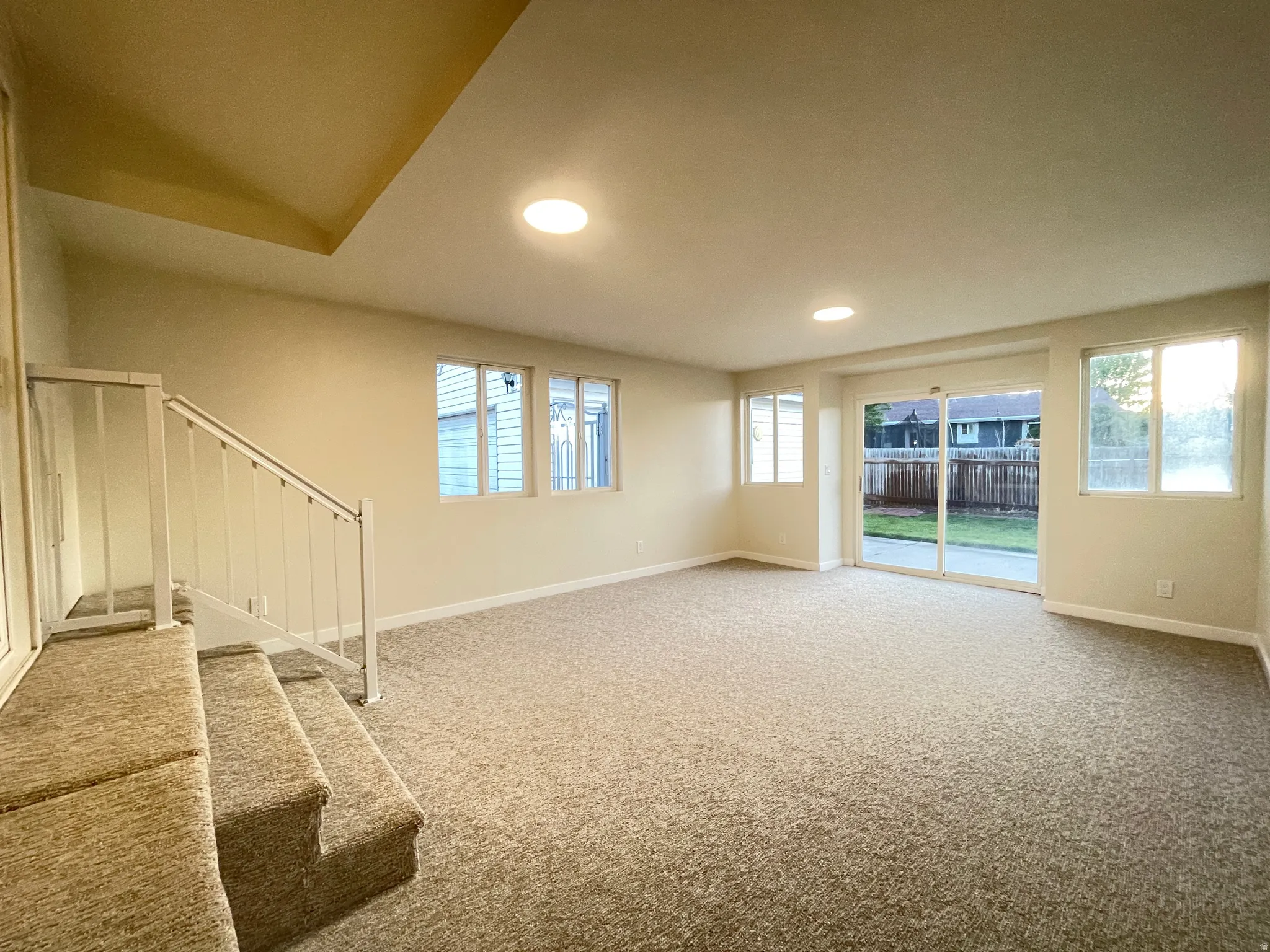 Spacious carpeted room featuring a white painted staircase with railing, multiple windows providing ample natural light, a sliding glass door offering backyard access, and recessed ceiling lighting