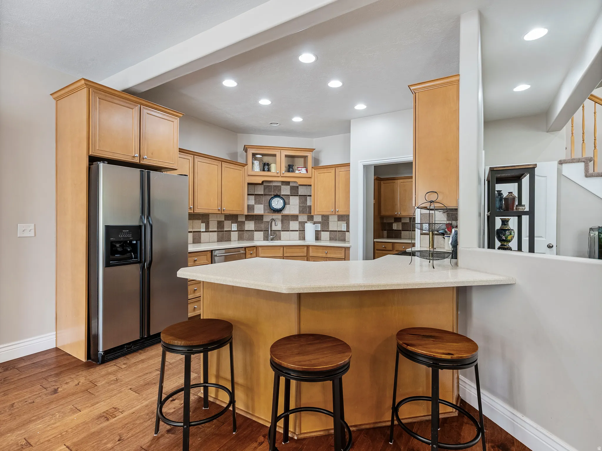 Kitchen featuring wood-finish flooring, light wood cabinetry, stainless steel refrigerator, recessed lighting, and a multi-toned tile backsplash