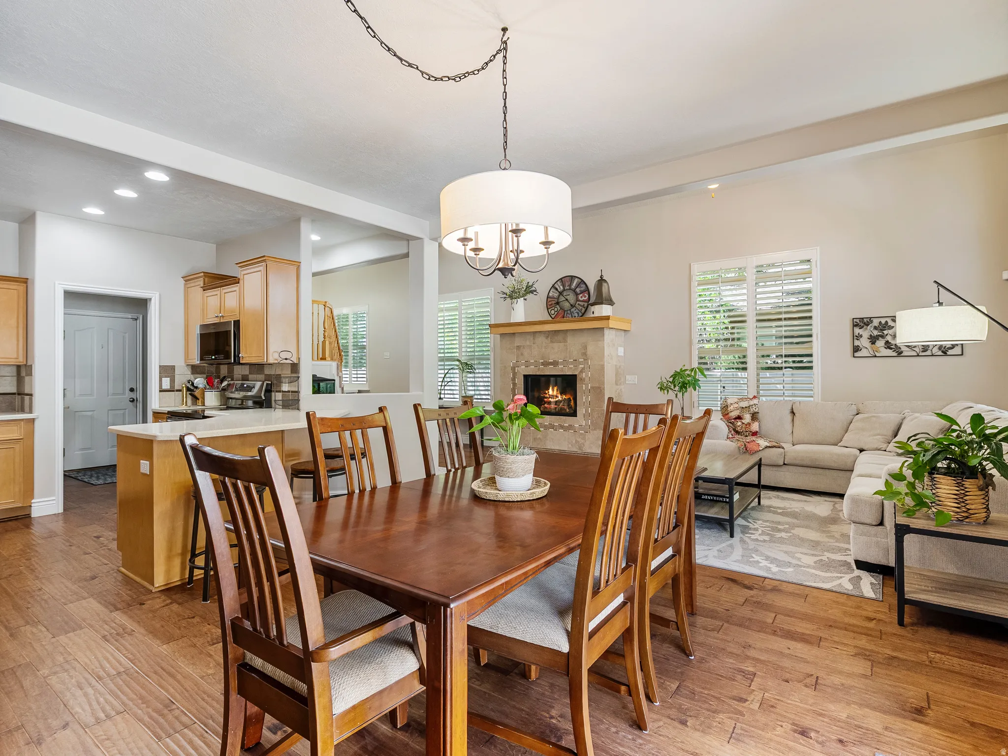 Open-concept living space featuring wood-finish flooring, a tiled fireplace with a wood mantel, and large windows with plantation shutters