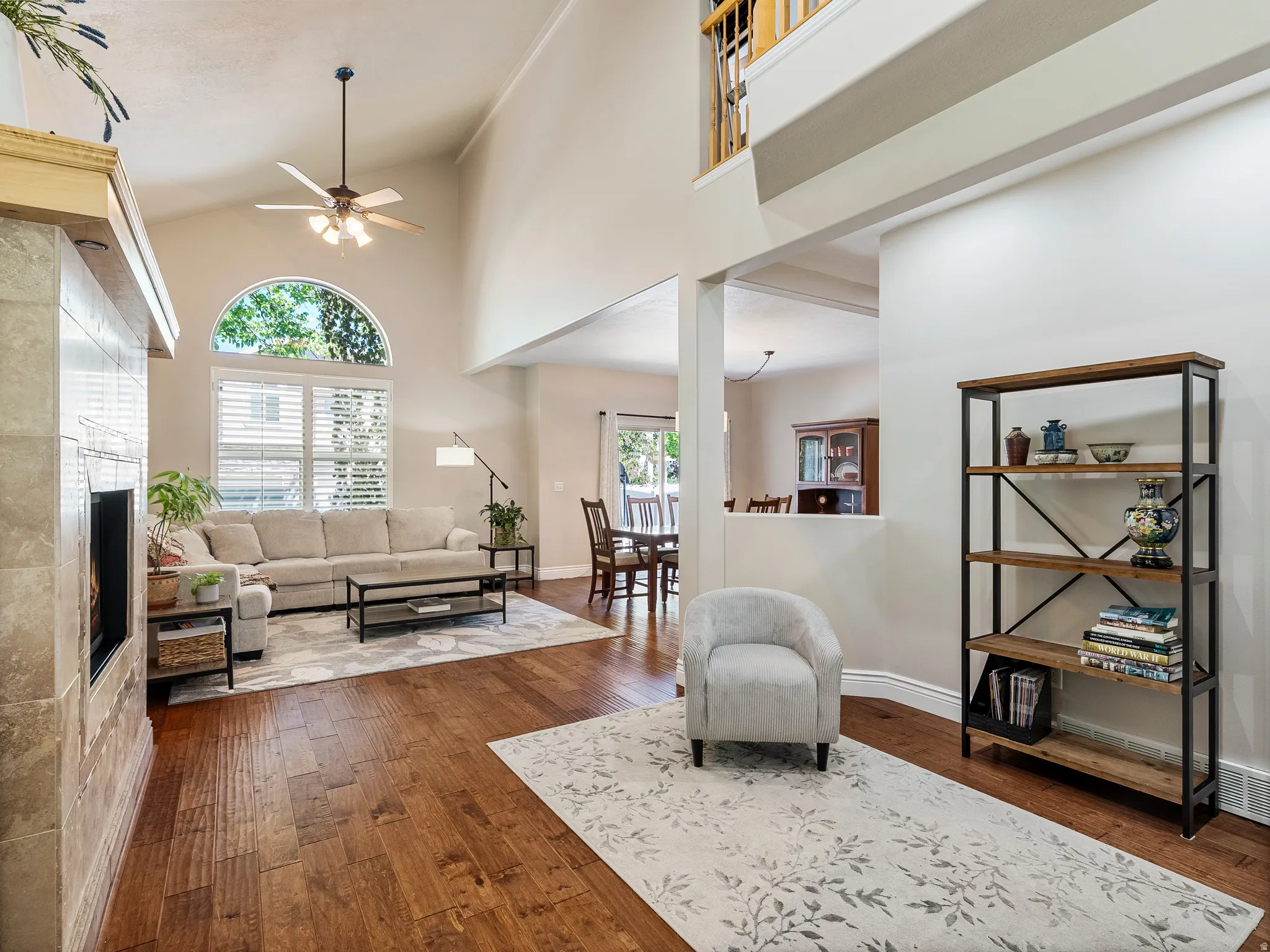 Spacious living area featuring vaulted ceilings, wood-finish flooring, a tile-surround fireplace, and a large arched window with plantation shutters