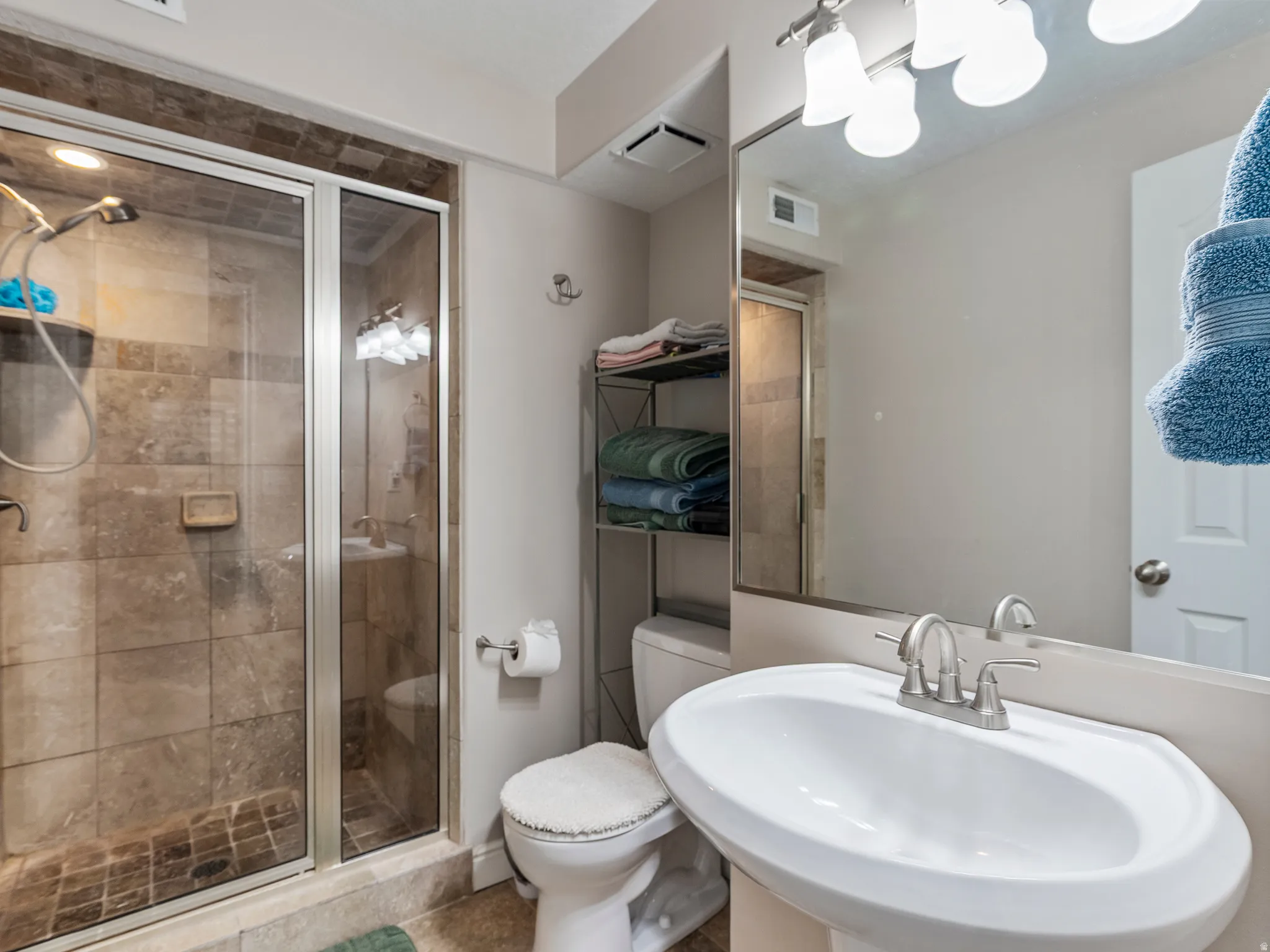 Bathroom featuring a glass-enclosed shower with stone-tile surround, a pedestal sink with a brushed nickel faucet, a large vanity mirror, and a wall-mounted light fixture