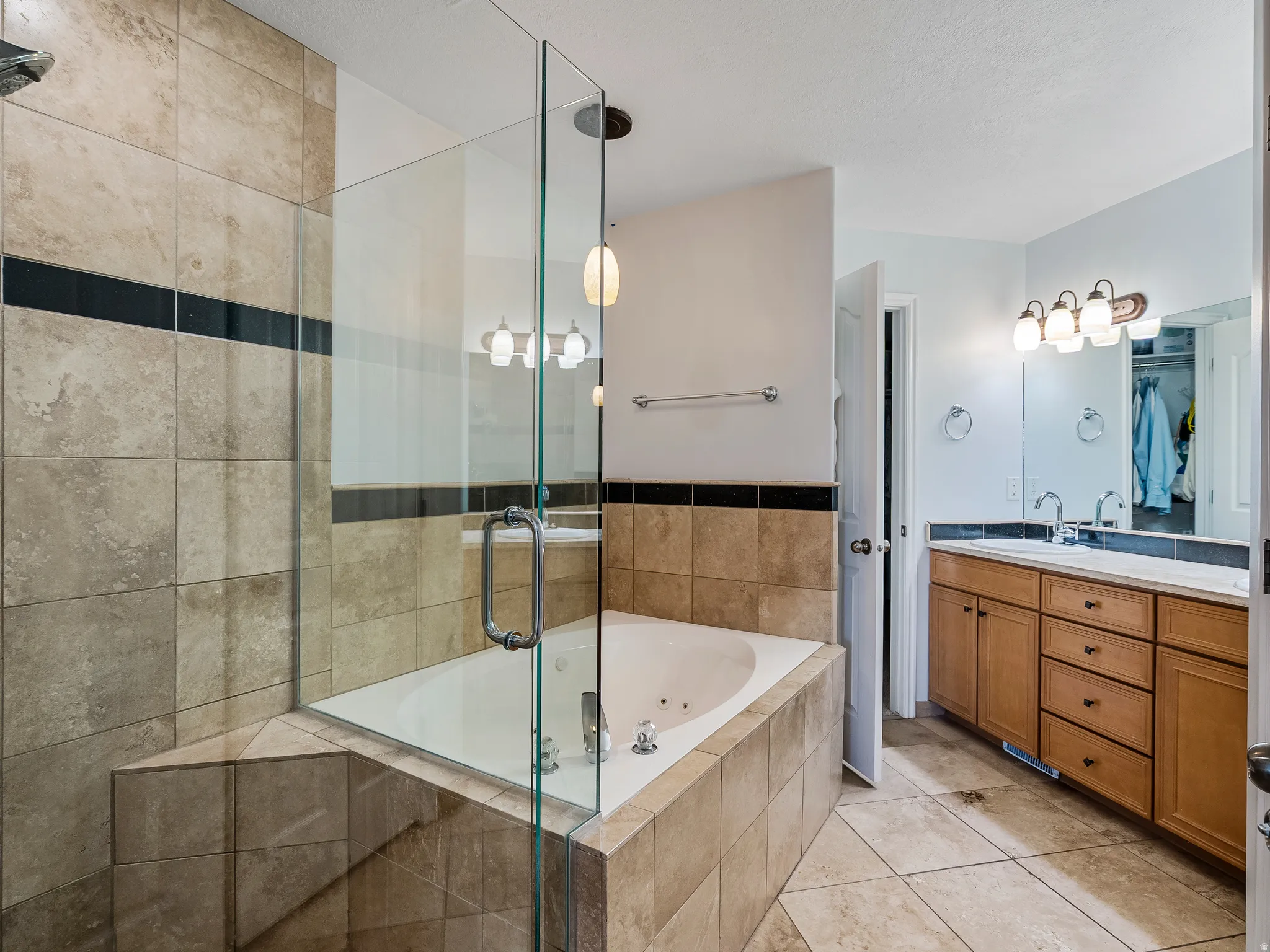 Tiled bathroom featuring a glass-enclosed shower, built-in corner tub with jets, and a double vanity with light wood cabinetry and dual sinks