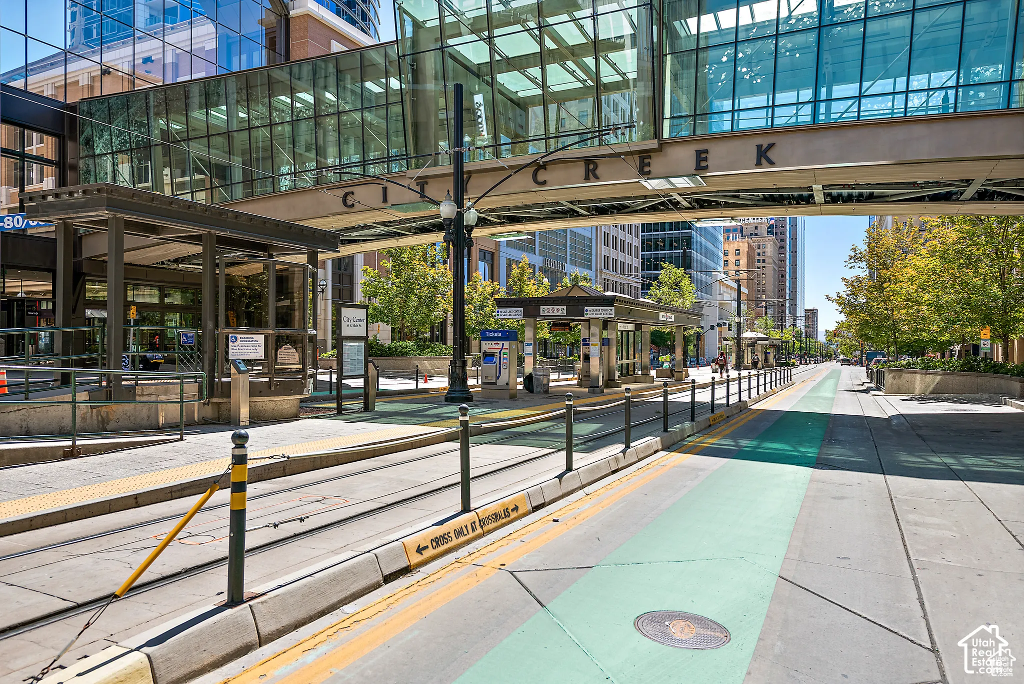 Urban street scene featuring light rail tracks, pedestrian walkways, and a covered station