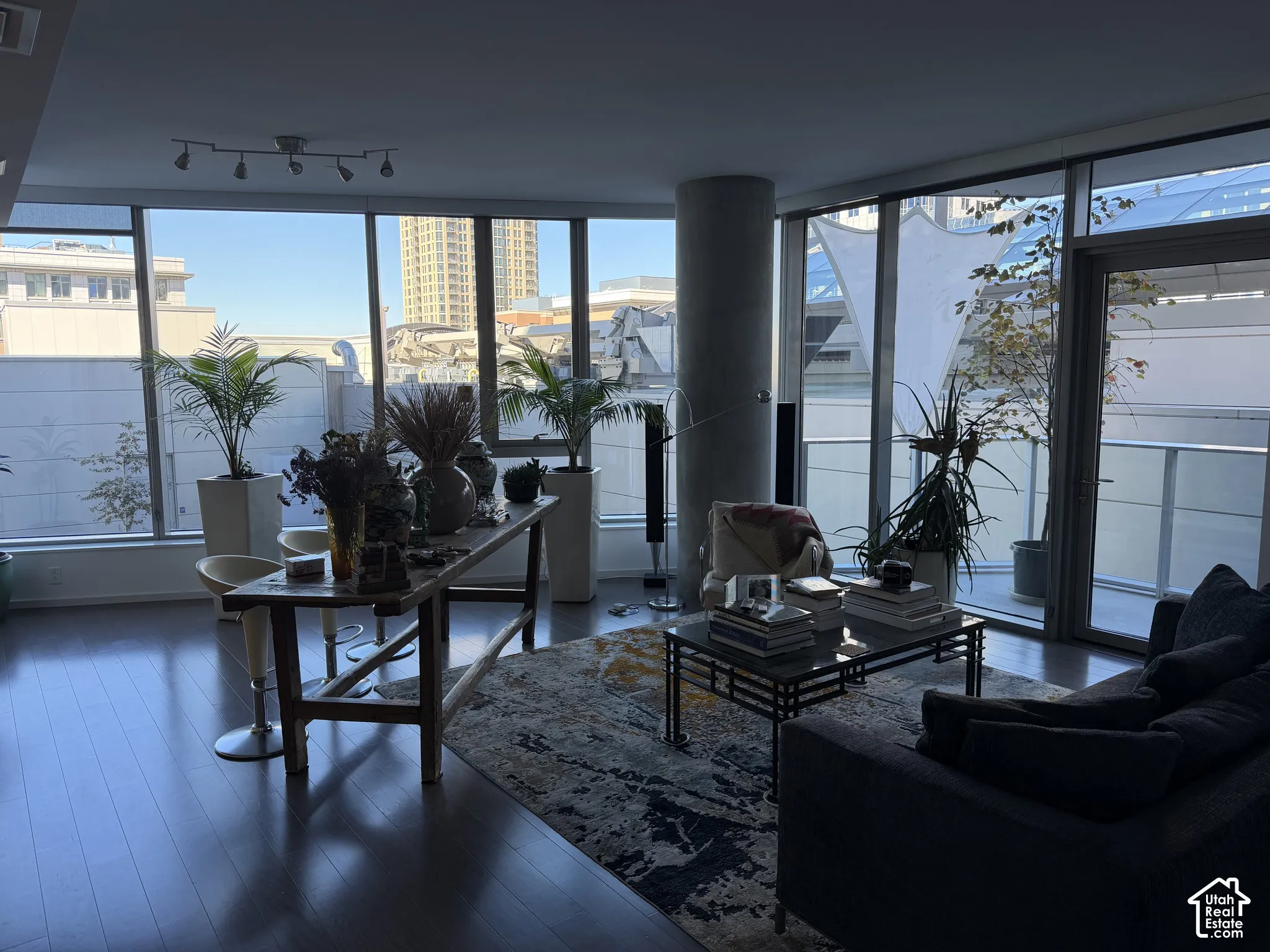 Expansive floor-to-ceiling windows, wood-finish flooring, concrete structural column, track lighting fixture, and a glass-paneled balcony