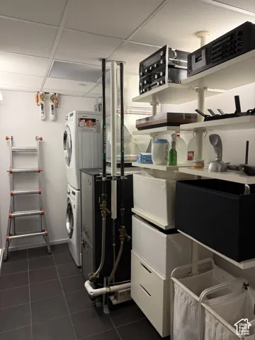 Laundry area featuring a stacked washer and dryer, dark-toned tile flooring, and a drop ceiling
