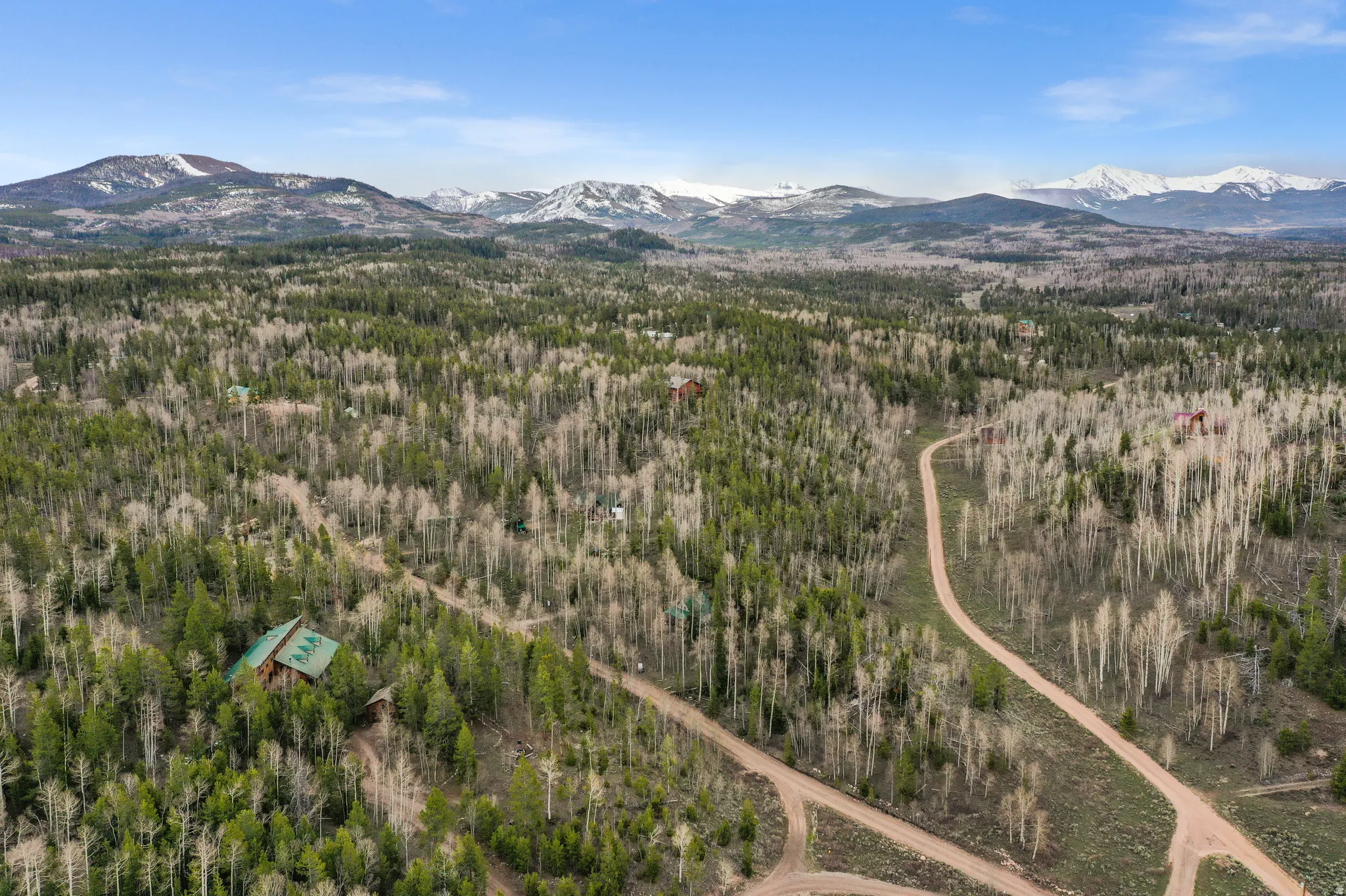 Expansive wooded property featuring a prominent main residence with a green metal roof, surrounded by mature evergreen and deciduous trees, and framed by distant snow-capped mountains