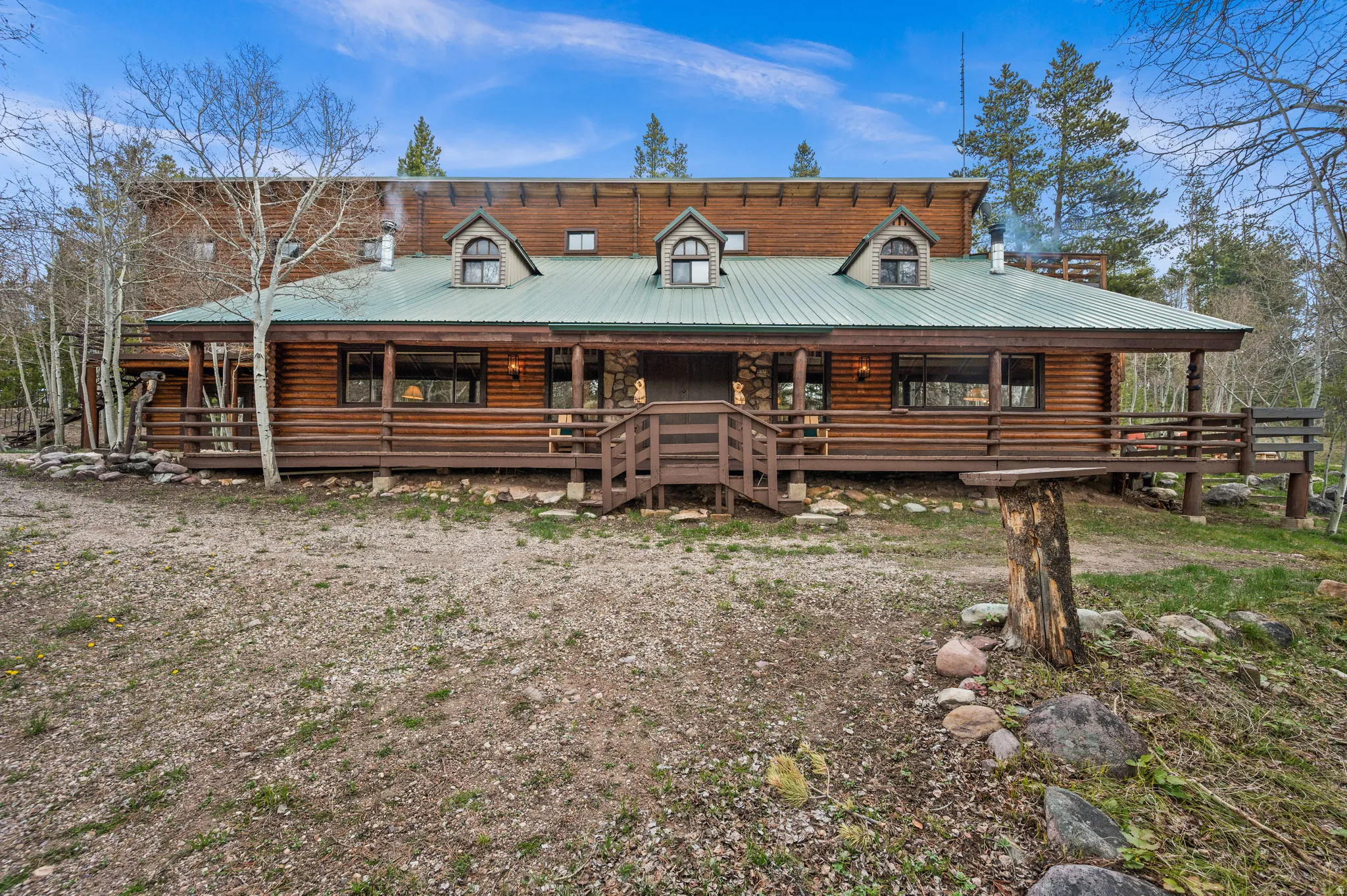 Log cabin exterior featuring a green metal roof, a full-length covered porch, multiple dormer windows, and a raised wooden deck with railings