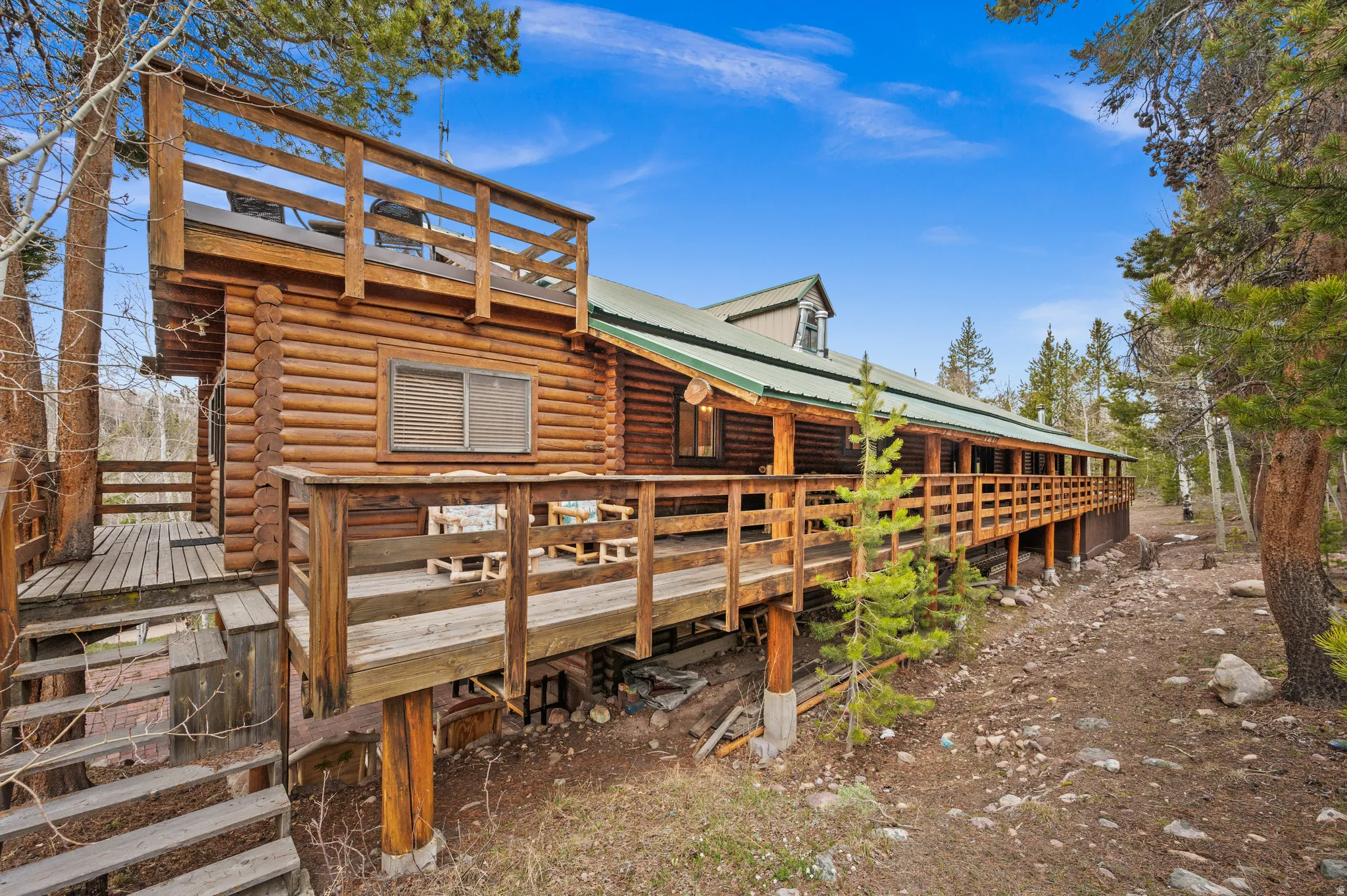Log cabin exterior featuring a second-story balcony, a wrap-around wooden deck, a prominent dormer, a metal roof, and a woodland setting