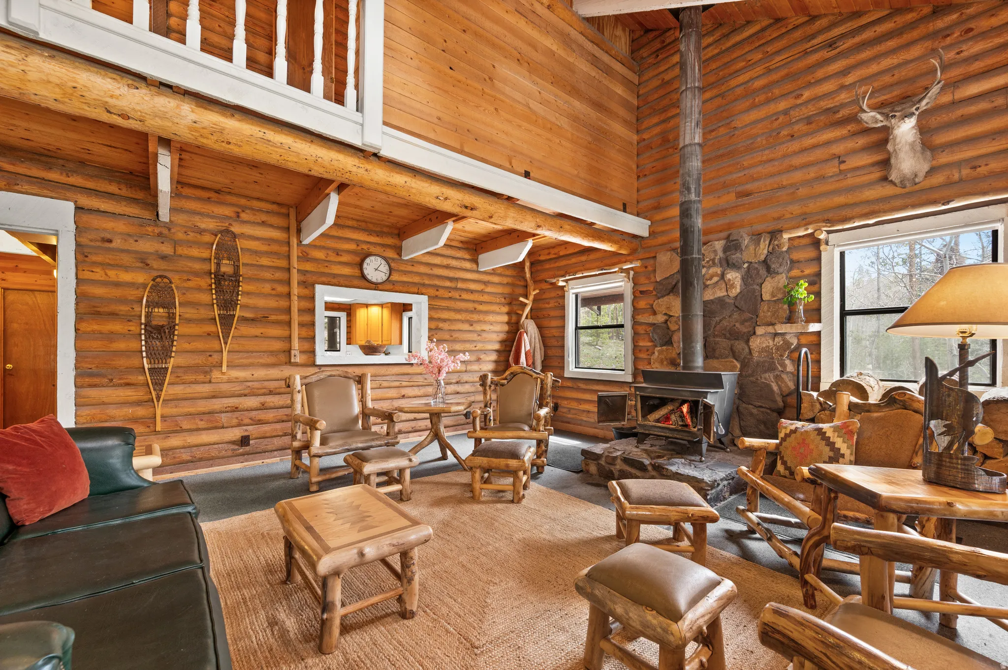 Expansive living area showcasing wood-finish log walls and ceilings, a prominent stone fireplace with a wood-burning stove, large windows, and a carpeted floor