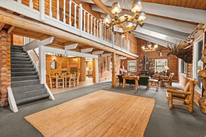 Expansive great room featuring a grand staircase with carpeted treads, a wood-burning stove with stone surround, and an upper-level balcony with white railings