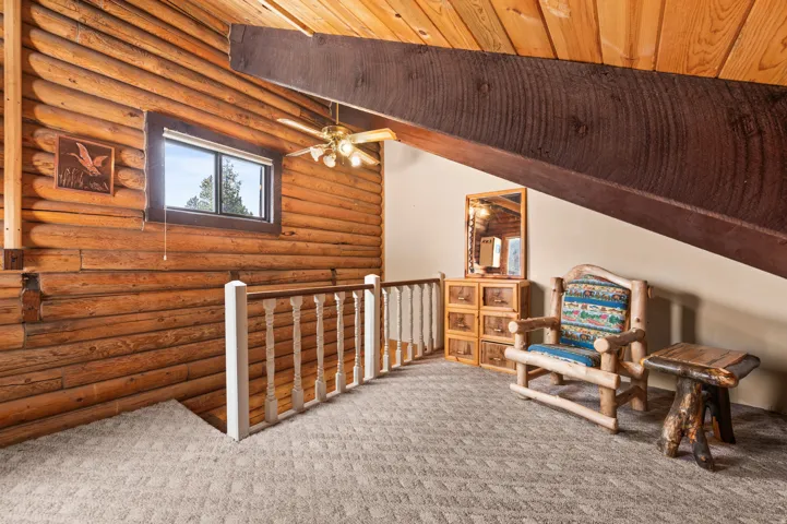 Loft area featuring log walls, an exposed wood beam ceiling, a ceiling fan with light fixture, and plush carpet flooring