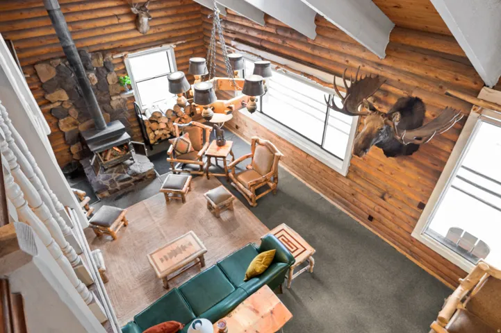 Spacious log cabin interior featuring a stone fireplace with wood stove, exposed log walls, high vaulted ceilings, and large windows