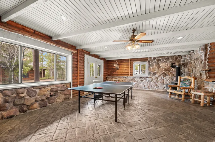 Spacious recreational room featuring a wood-planked ceiling with exposed beams, stamped concrete flooring, extensive window banks, rustic log walls, and a stone accent wall