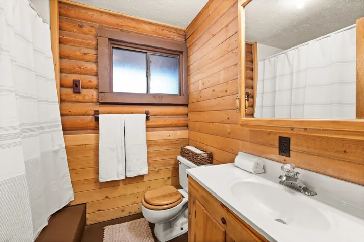 Bathroom featuring log and wood paneling, a white vanity with an integrated sink, a framed mirror, a toilet with a wooden seat, and a window with frosted glass