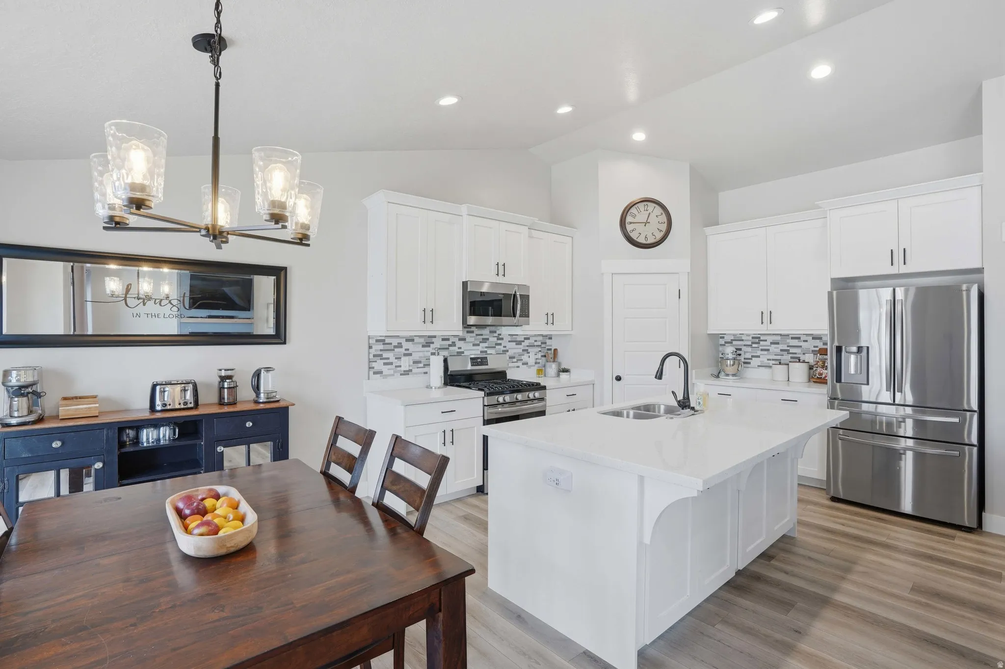 Contemporary kitchen and dining area featuring white shaker-style cabinetry, stainless steel appliances, a center island with sink, mosaic tile backsplash, and wood-finish flooring