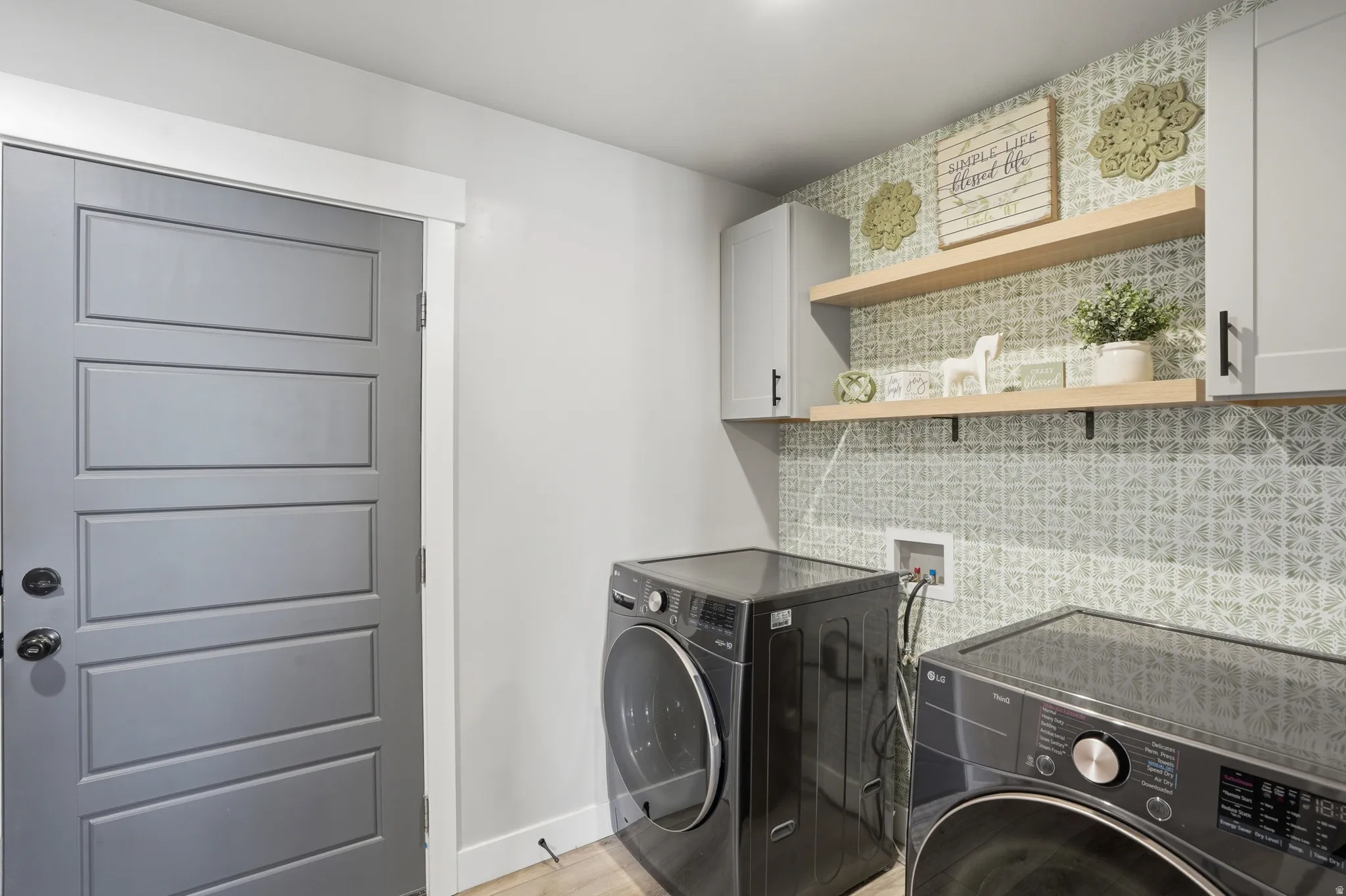 Laundry space featuring light grey cabinetry, natural wood-finish floating shelves, and a patterned wall covering