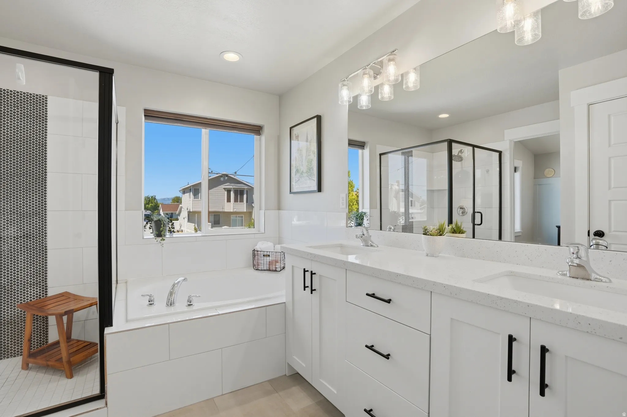 Primary bedroom with dual vanity with white cabinetry and quartz countertops, featuring a built-in soaking tub, a separate glass-enclosed shower with black-framed accents, and tile flooring