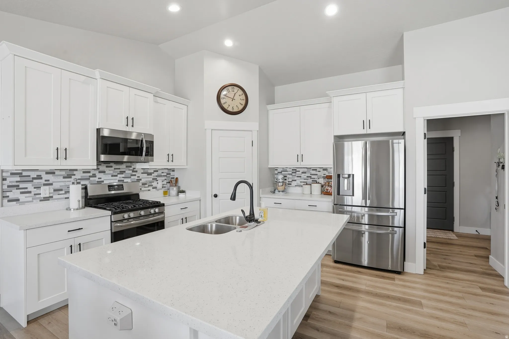 Kitchen featuring white cabinetry, stainless steel appliances, a large central island with a double basin sink, mosaic tile backsplash, and light wood-finish flooring
