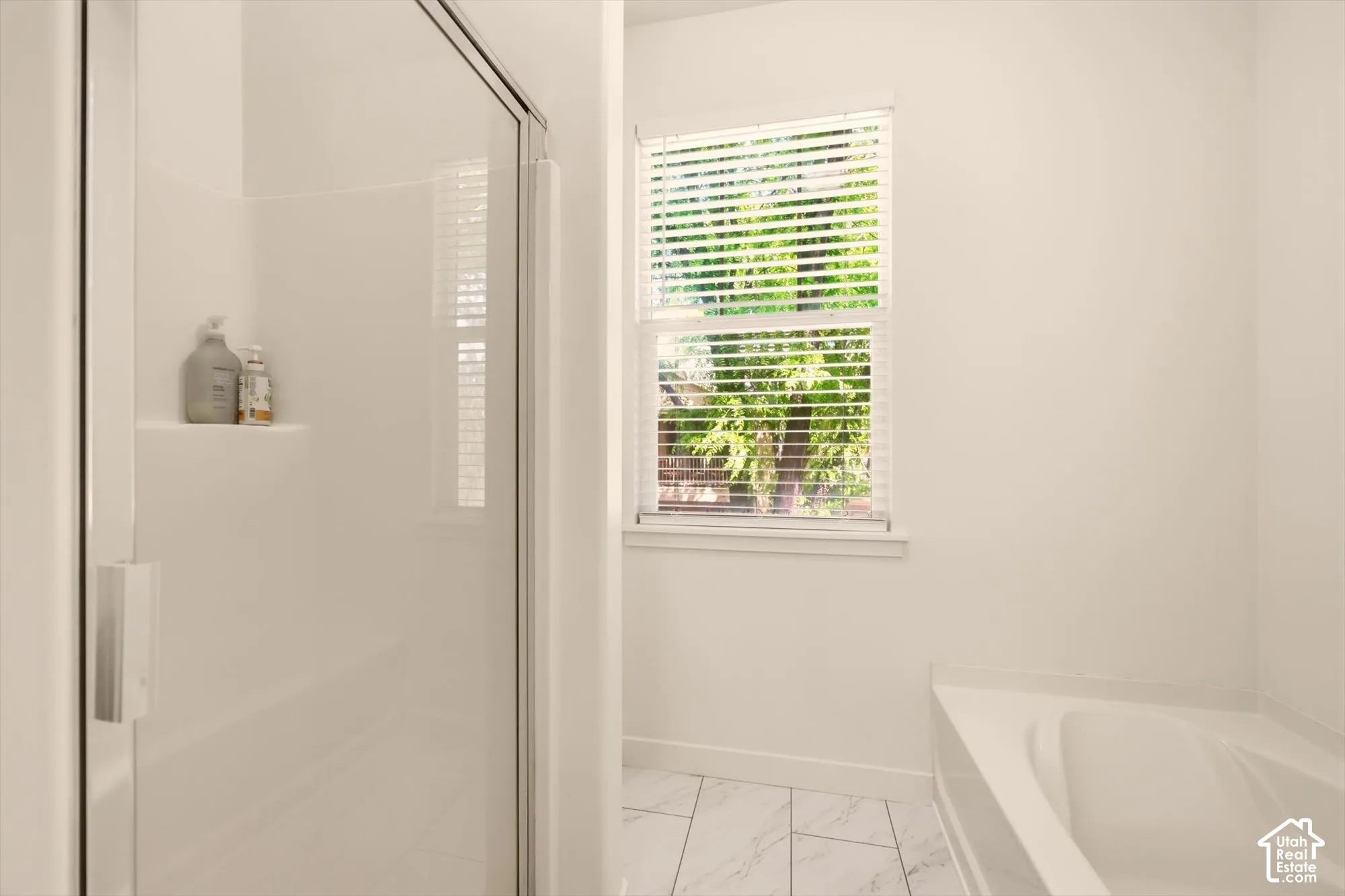White shower enclosure with clear glass door, built-in shelving, and an adjacent window with blinds above a built-in bathtub