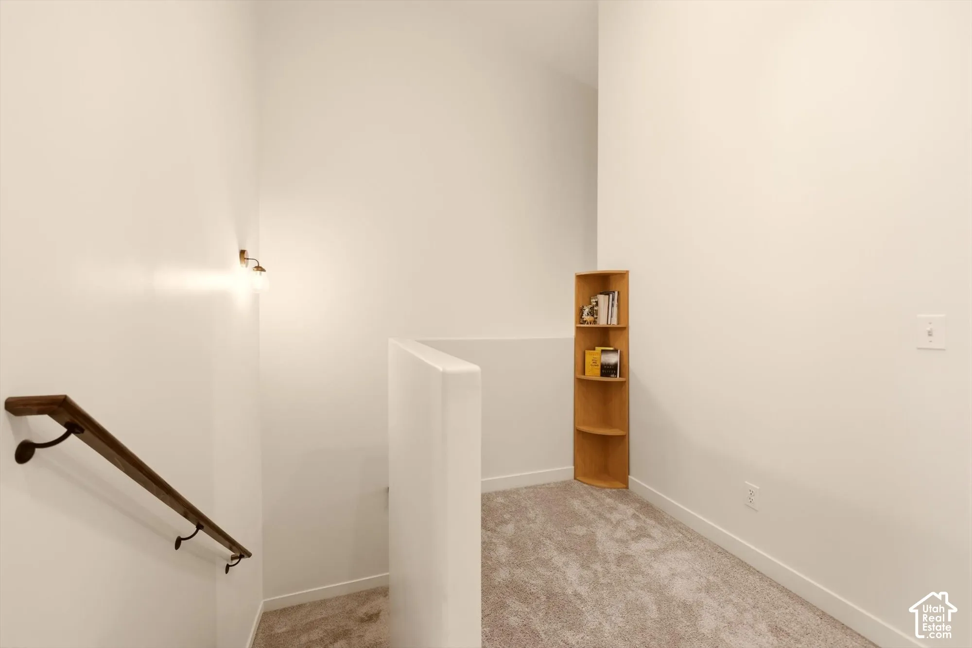 Carpeted landing area featuring a wood handrail with dark metal brackets, bright white walls, and a built-in wood corner shelf
