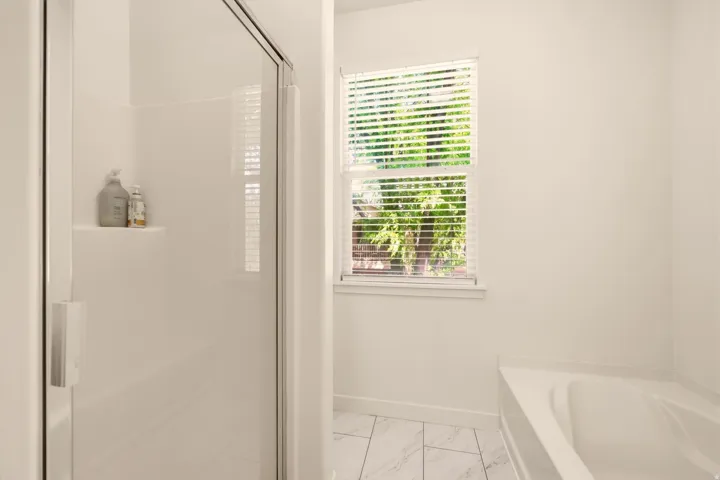 White shower enclosure with clear glass door, built-in shelving, and an adjacent window with blinds above a built-in bathtub
