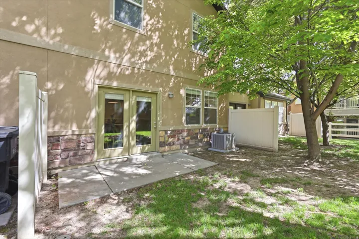 Ground-level patio with double glass doors, stucco and stone exterior, mature shade trees, and partial privacy fencing