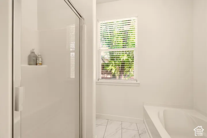 White shower enclosure with clear glass door, built-in shelving, and an adjacent window with blinds above a built-in bathtub