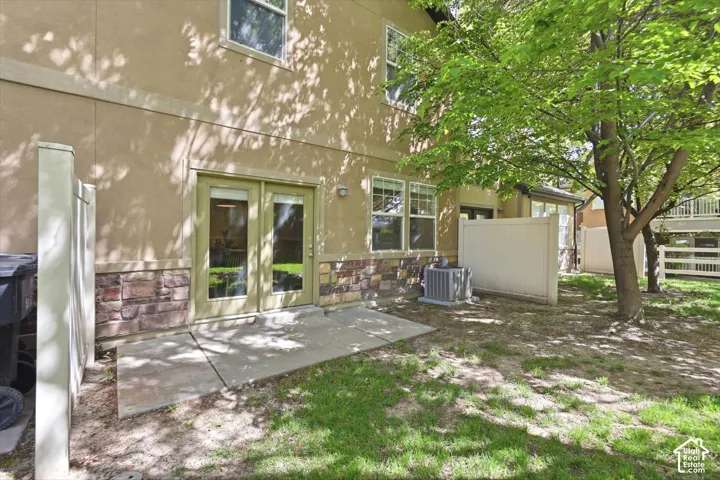Ground-level patio with double glass doors, stucco and stone exterior, mature shade trees, and partial privacy fencing