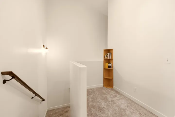 Carpeted landing area featuring a wood handrail with dark metal brackets, bright white walls, and a built-in wood corner shelf