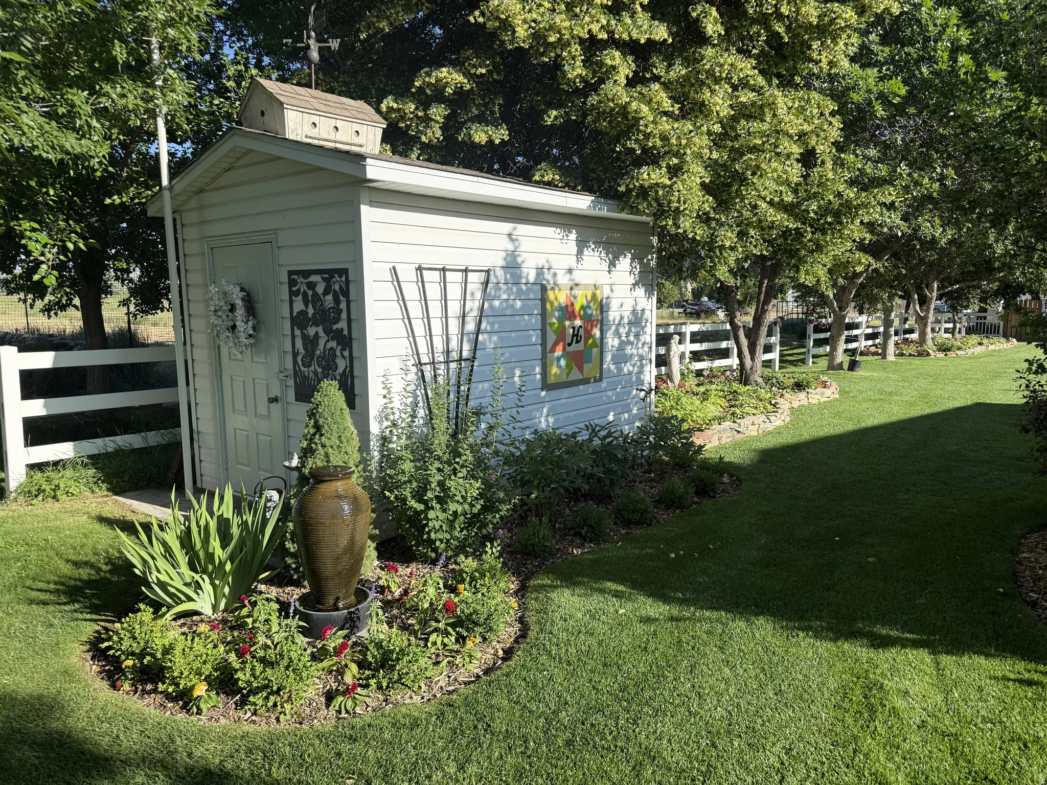 Expansive lawn area featuring a white vinyl-sided shed with a birdhouse and weathervane, bordered by a white picket fence