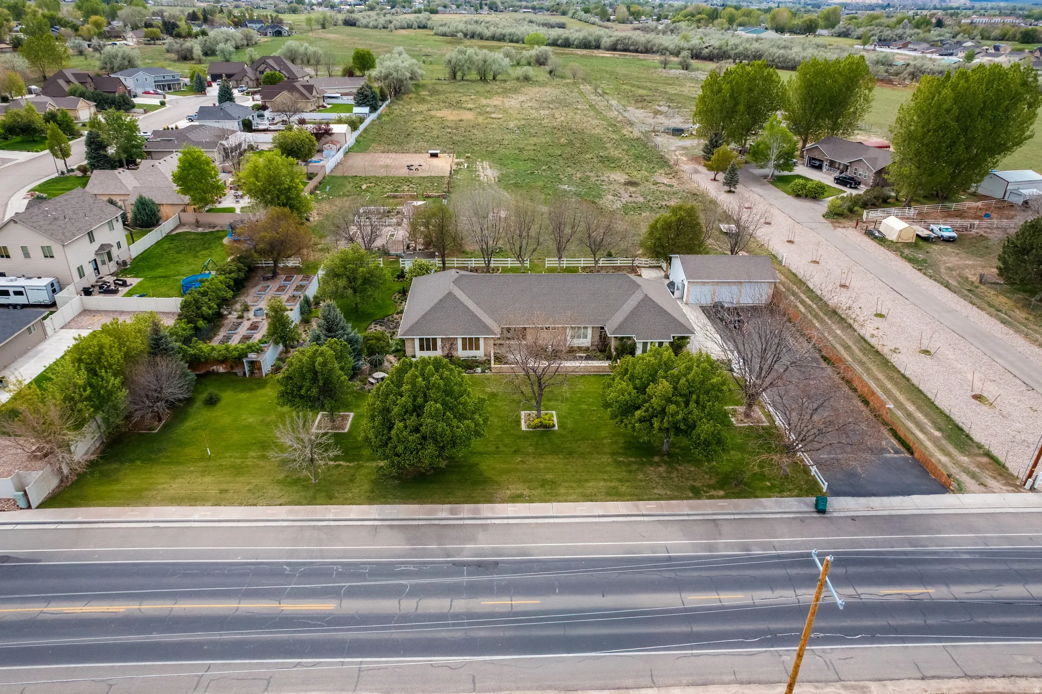 Ranch-style residence featuring a prominent front lawn with mature trees, an expansive cultivated garden area, an attached two-car garage, and an additional detached outbuilding