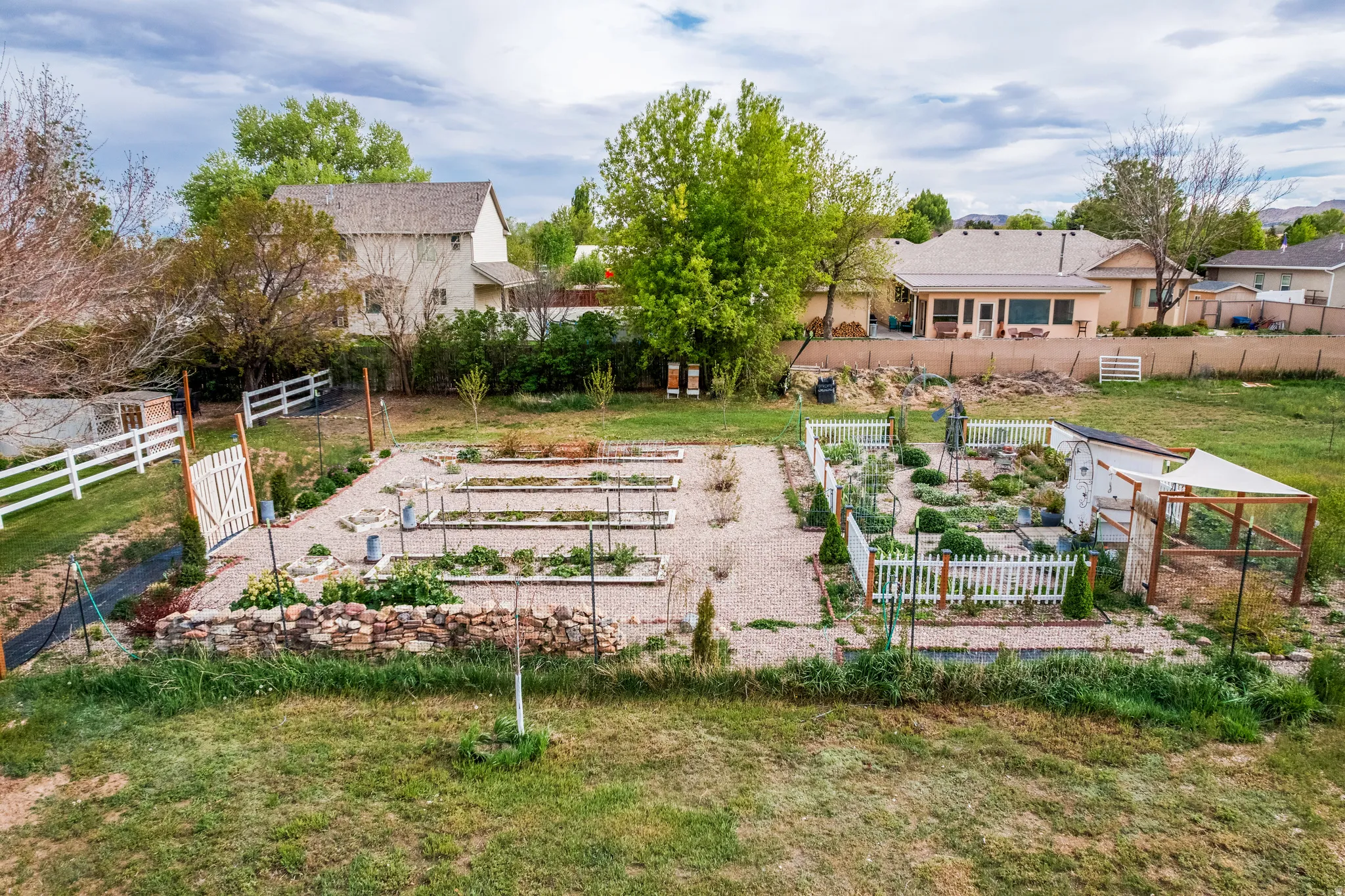 Expansive rear yard featuring multiple garden beds with diverse plantings, white picket fencing, and a stone retaining wall