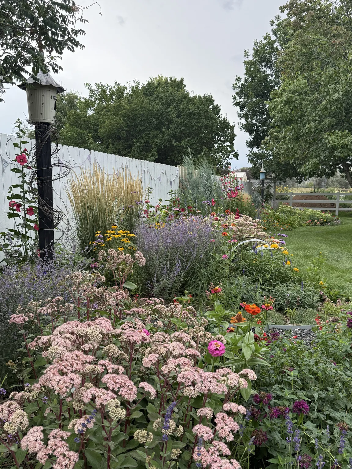Expansive garden bed featuring diverse plantings, a white picket fence, and mature perimeter trees