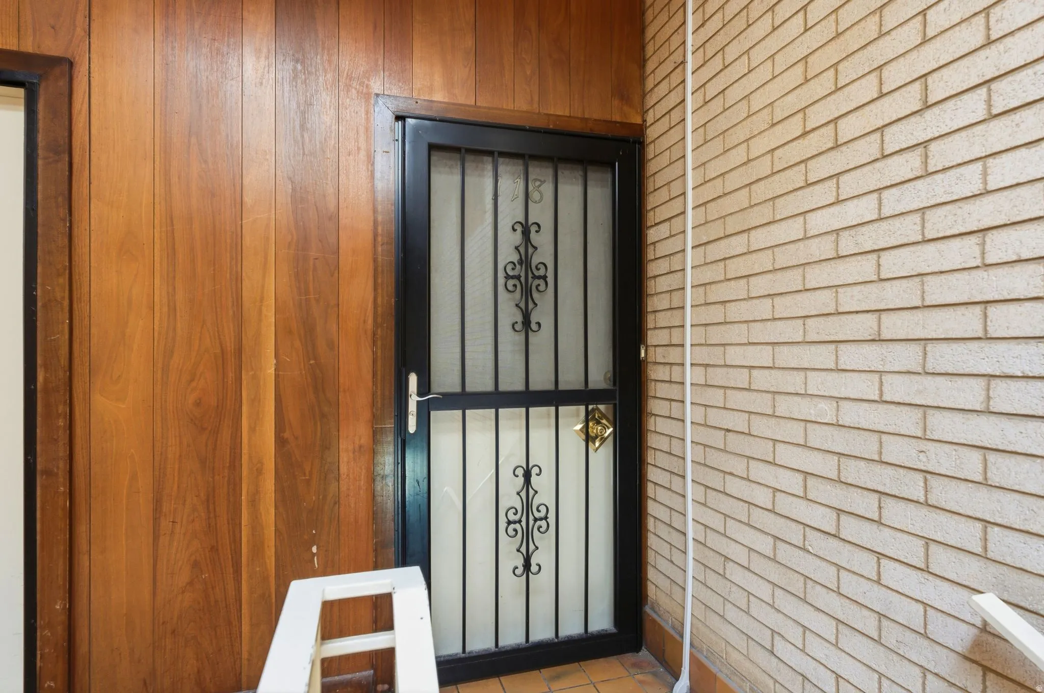 Black wrought iron security screen door with decorative scrollwork, set against warm wood-paneling and a light-toned brick wall