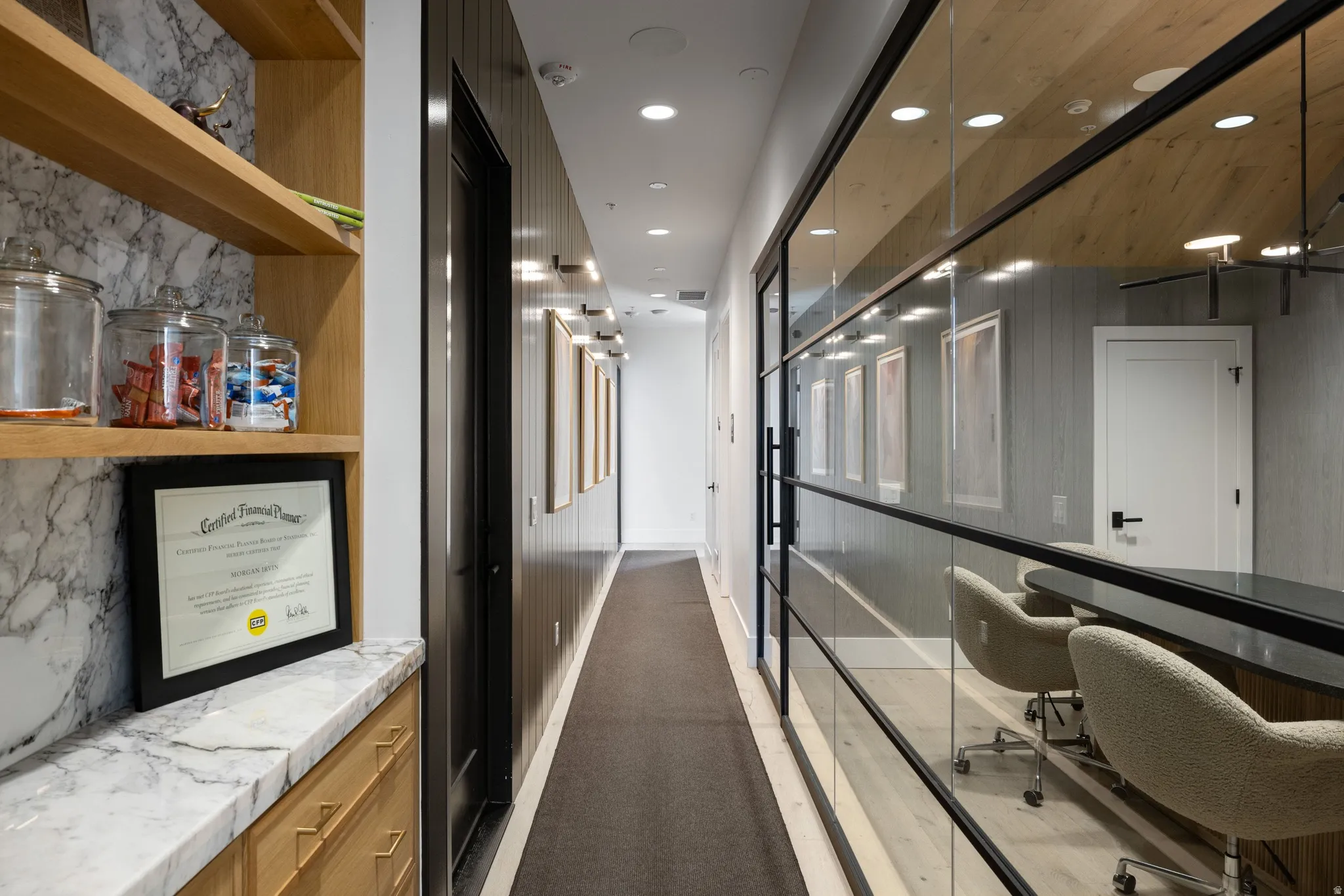 Hallway featuring a glass wall with black framing, wood-finish wall panels, recessed lighting, and a built-in storage unit with marble-finish countertop and light wood shelving