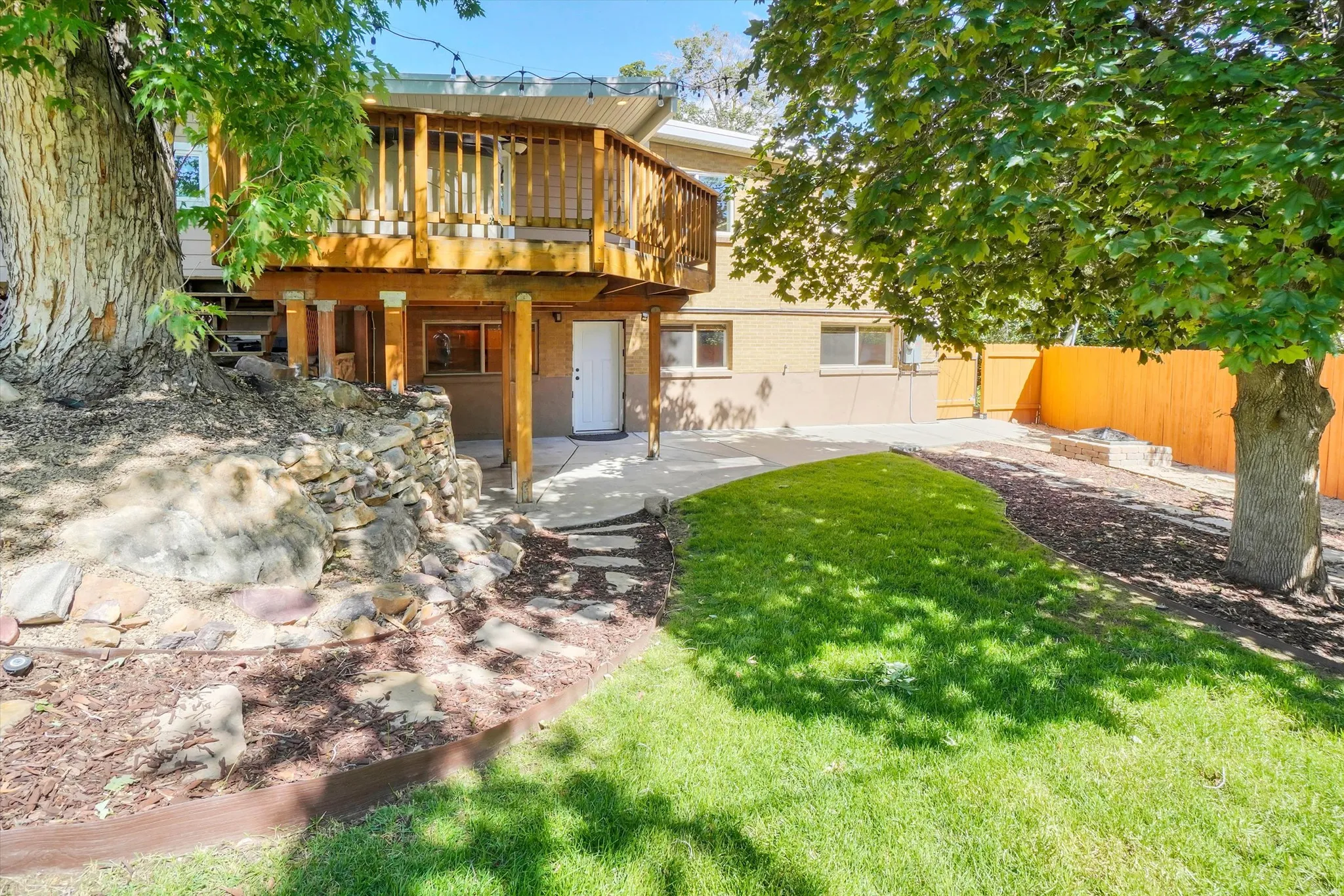 Rear exterior featuring a spacious wood deck, lower-level patio, mature trees, and a manicured lawn area