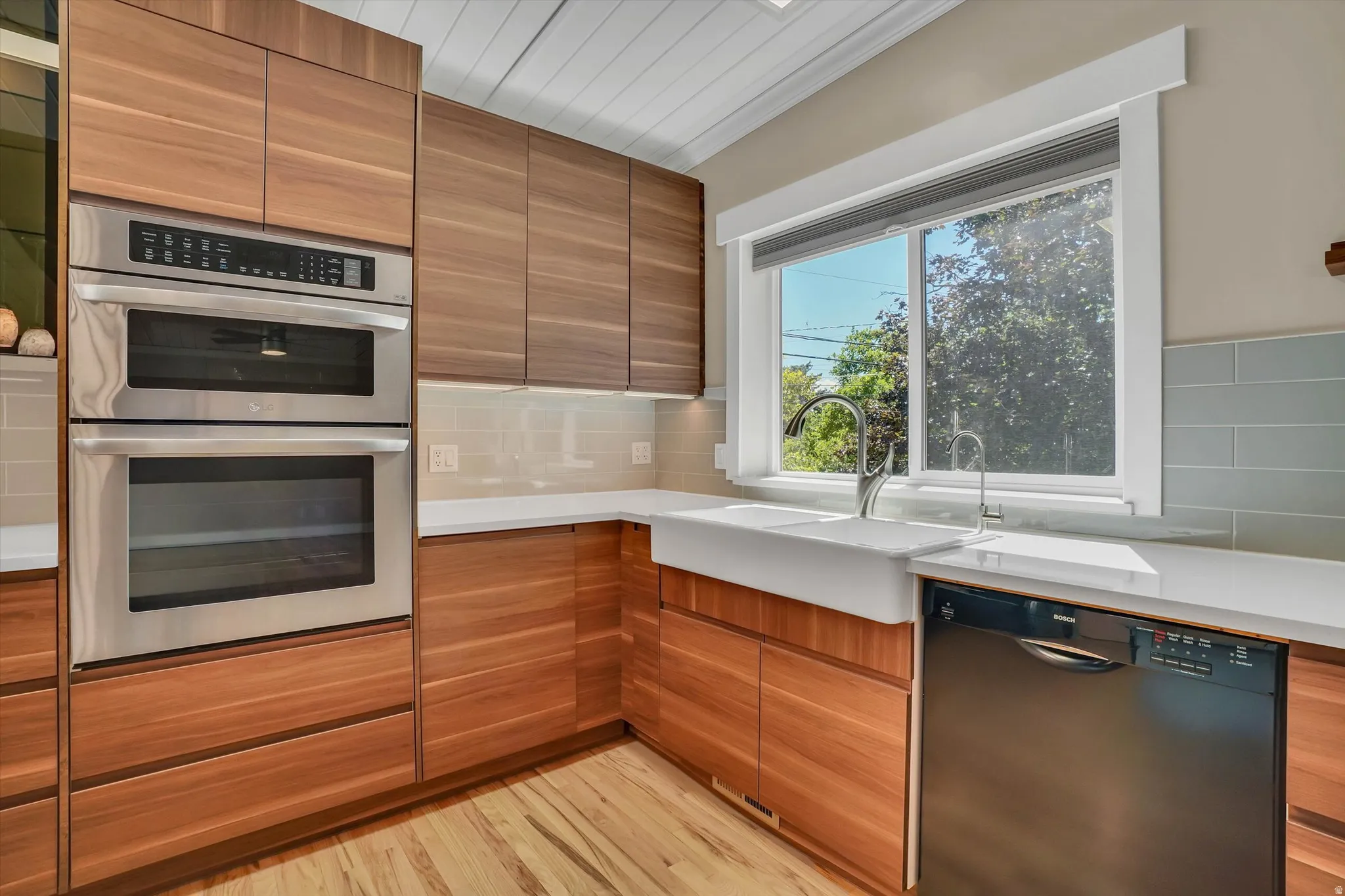 Kitchen featuring wood-finish cabinetry, stainless steel double wall ovens, a large farmhouse sink, a dishwasher, and light countertops