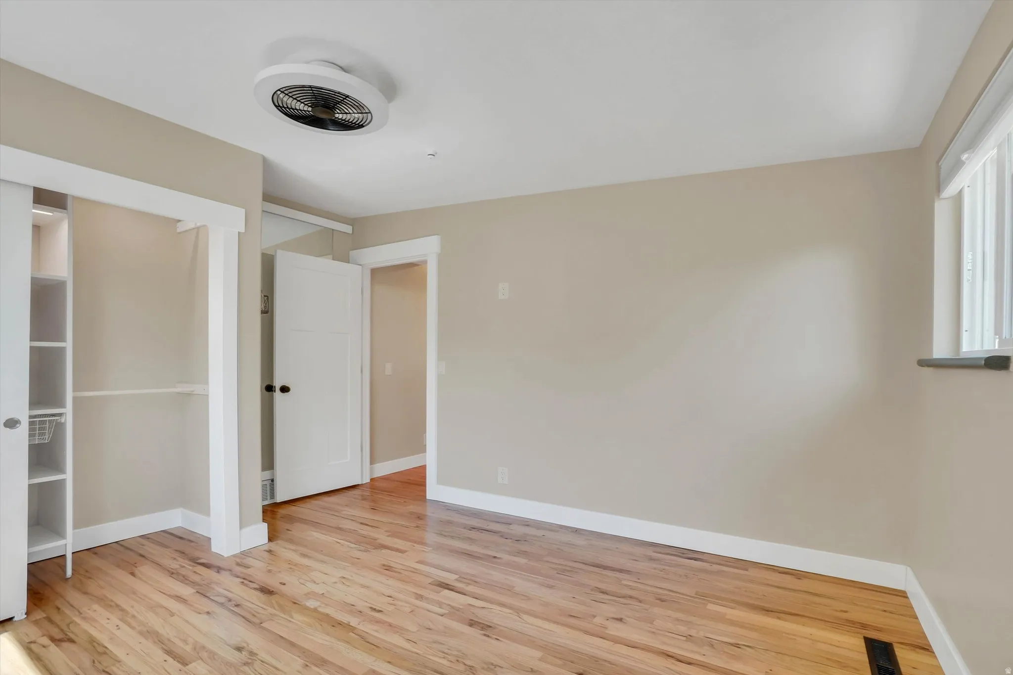 Room featuring wood-finish flooring, a built-in closet system with shelving, a ceiling-mounted fan, a window with blinds, and a white paneled door