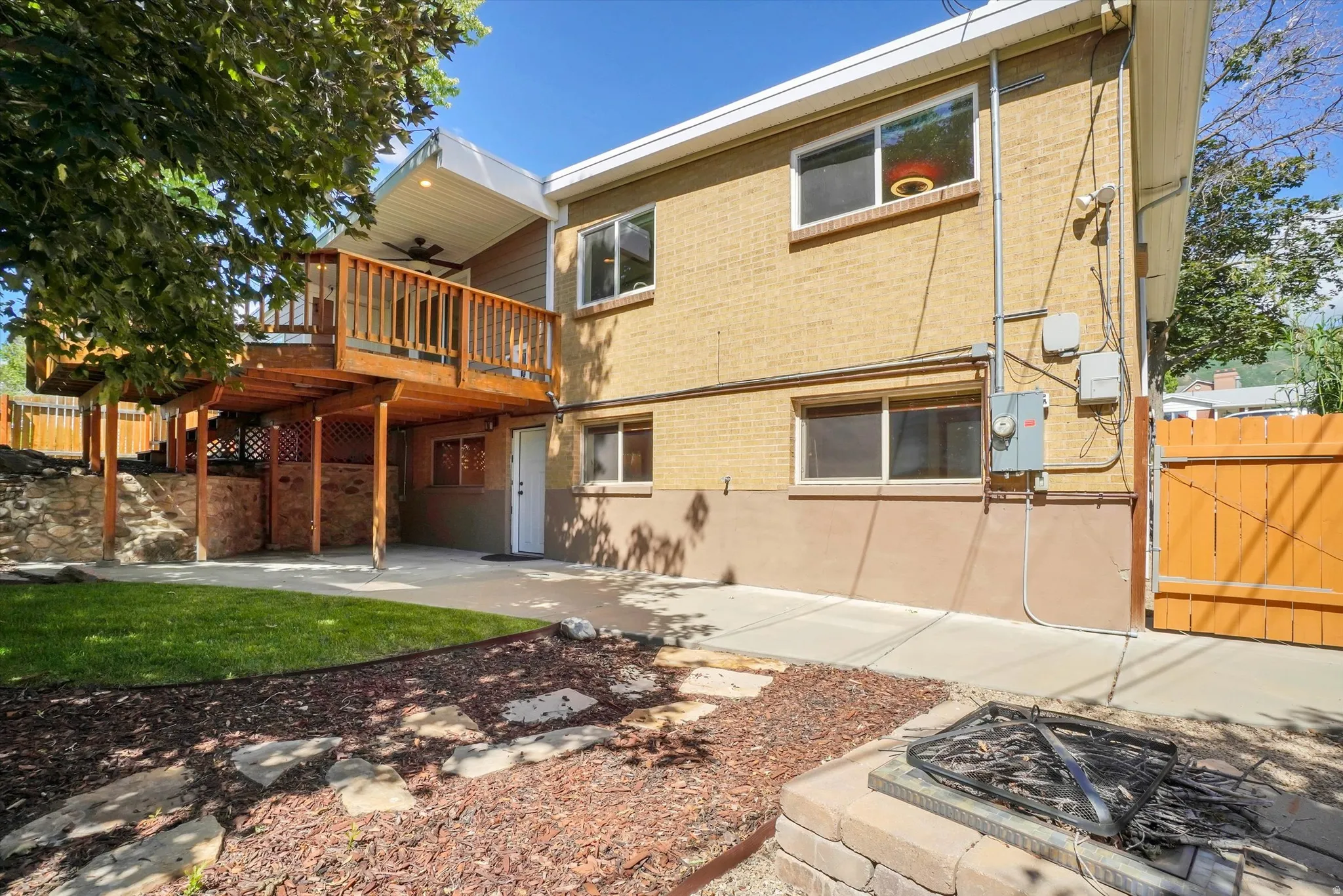 Rear exterior featuring a multi-level wood deck with a ceiling fan, a lower concrete patio, a built-in stone fire pit, a vibrant green lawn, and mature landscaping