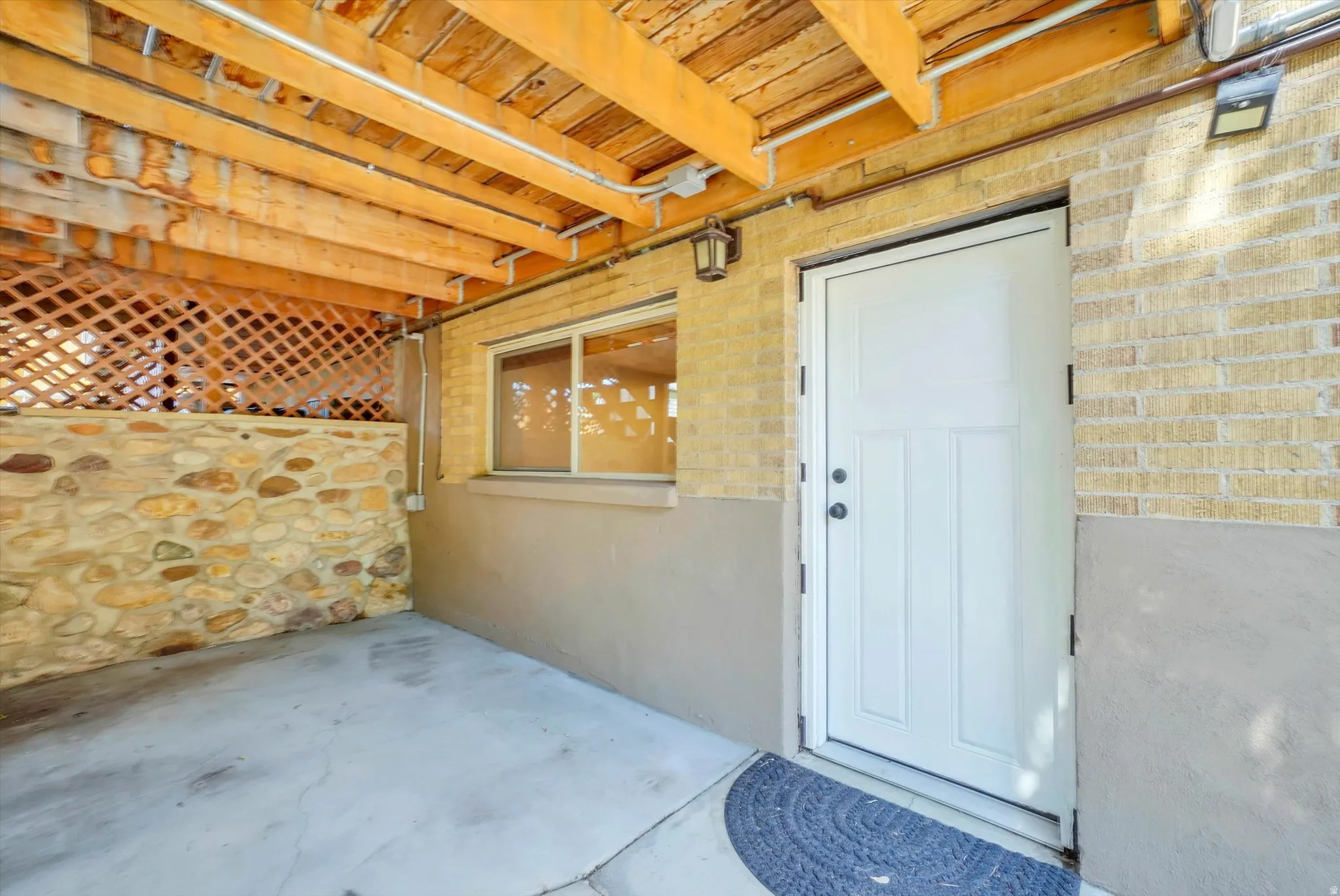 Outdoor covered patio area featuring a white entry door, an exterior wall with light-colored brick, a stone half-wall, wood lattice, and a wood beam ceiling structure