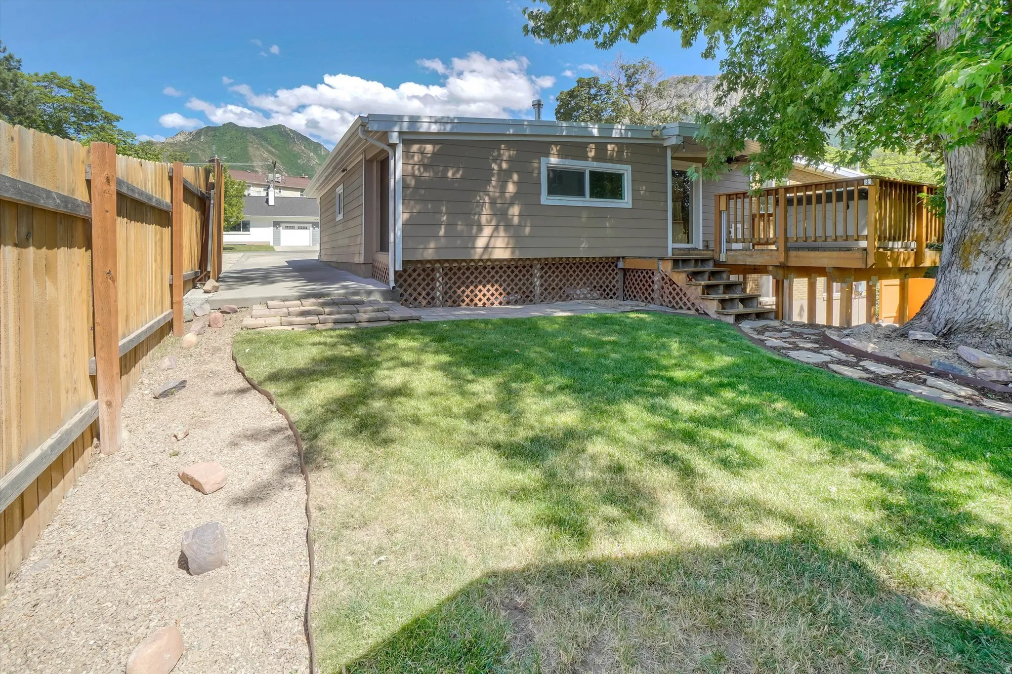 Expansive backyard featuring a manicured lawn, a wooden privacy fence, a gravel pathway with stone accents, a multi-level wooden deck, and mature trees