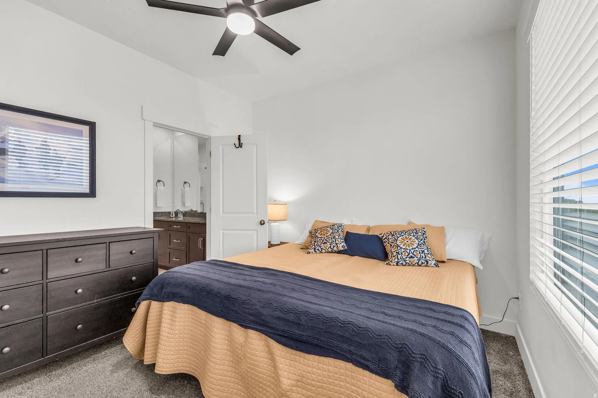 Carpeted bedroom featuring a ceiling fan with integrated lighting, a window with horizontal blinds, and an attached bathroom with a double vanity