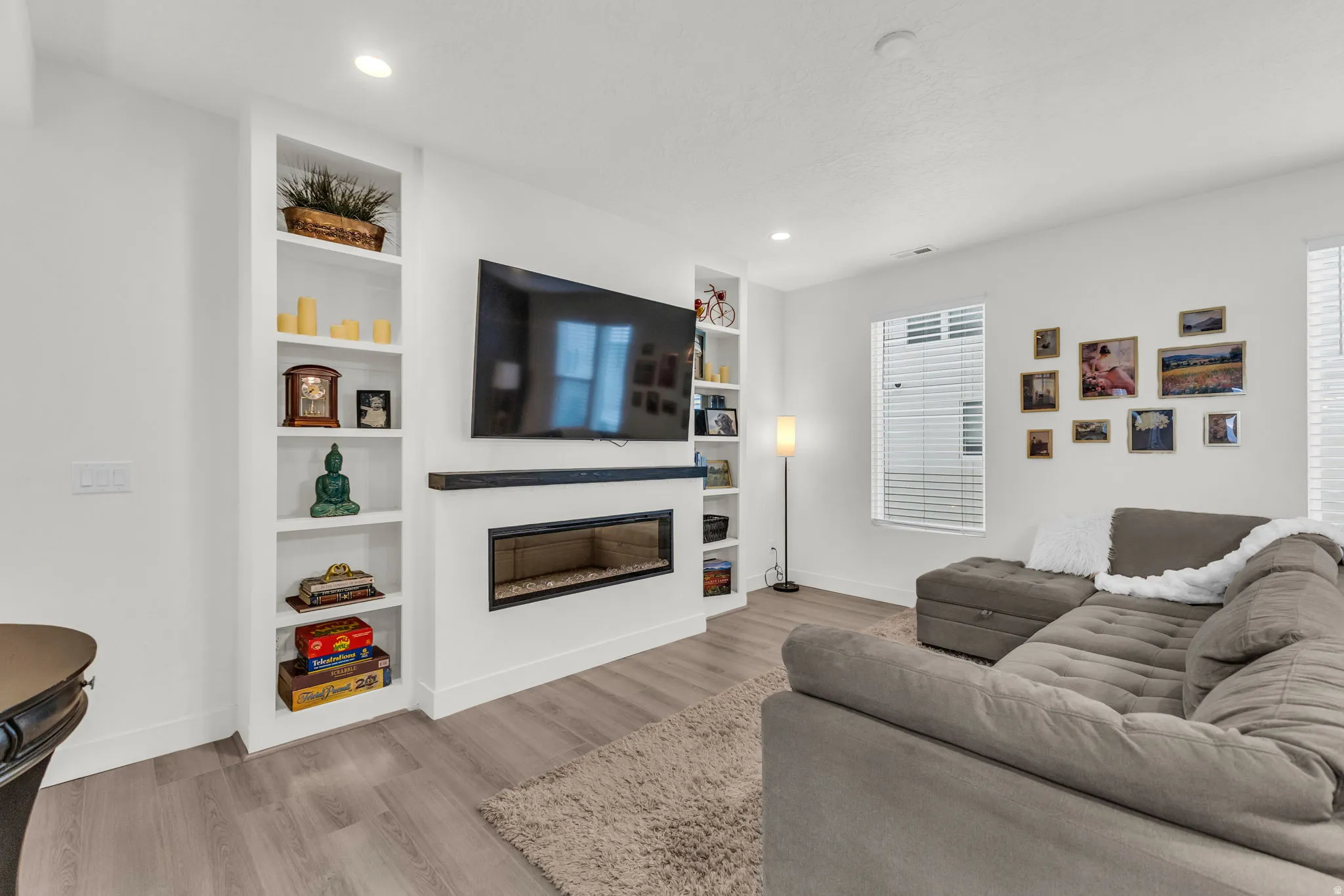 Living area featuring an integrated linear electric fireplace with a black wood mantel, built-in shelving, wood-finish flooring, recessed lighting, and white window blinds