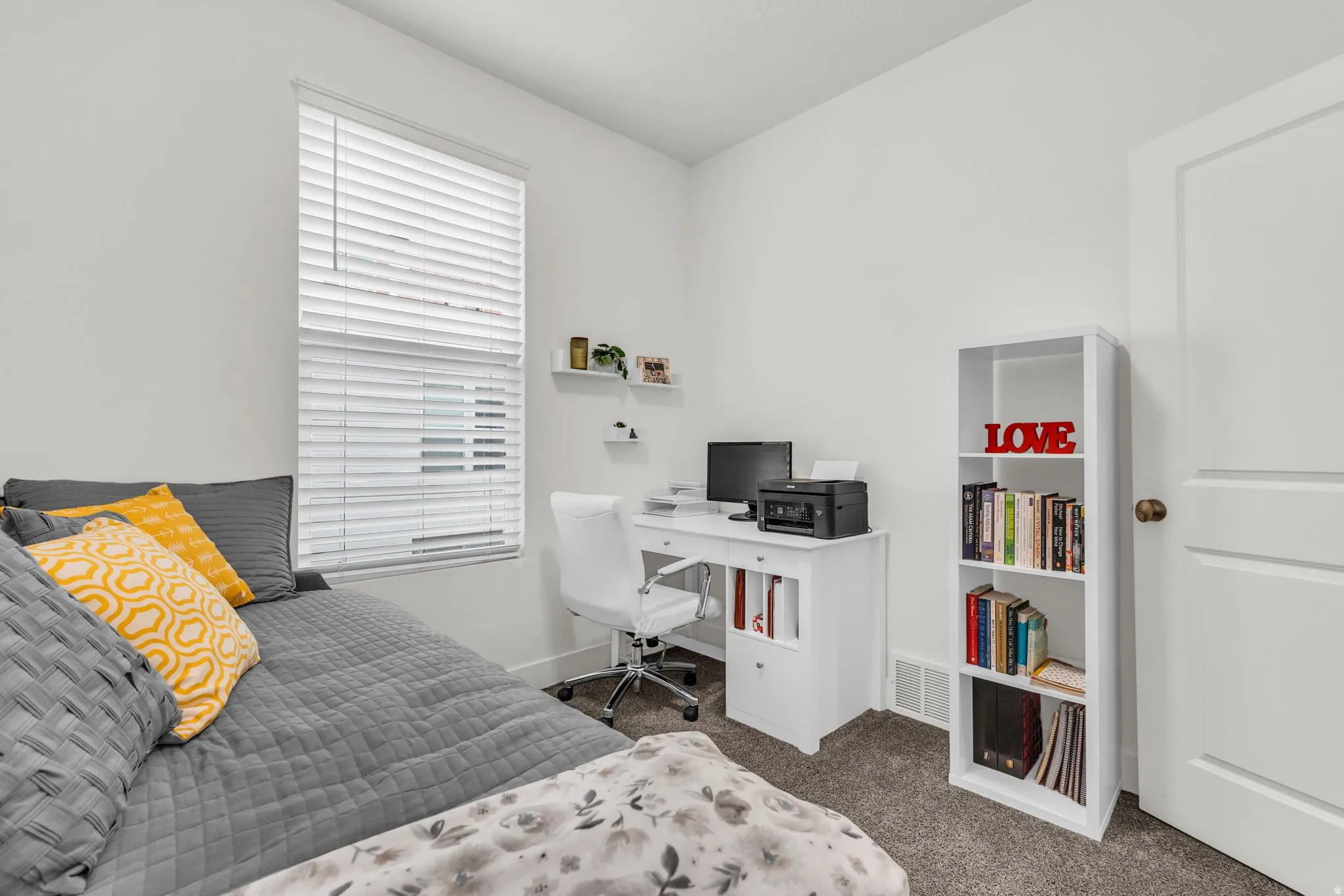 Light-filled room featuring a broad window with blinds, soft gray carpeting, white painted walls, a built-in white desk, and white floating shelves