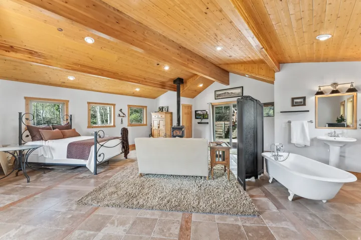 Vaulted wood-plank ceilings with exposed beams, wood-frame windows, a freestanding clawfoot tub, and a wood-burning stove