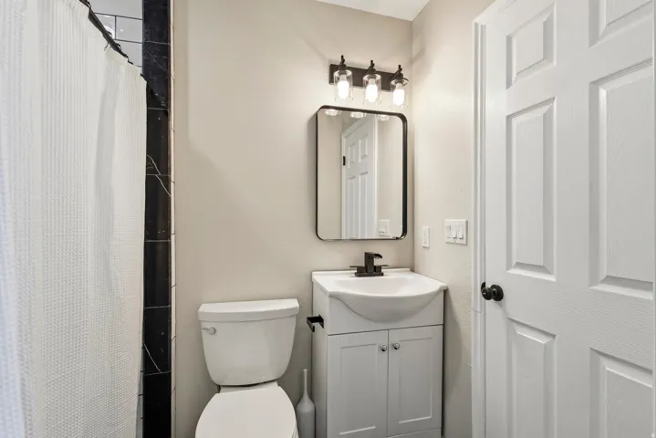 Compact bathroom featuring a white vanity with integrated sink, black metal-framed mirror, contemporary three-bulb vanity light, white toilet, and black tile shower surround