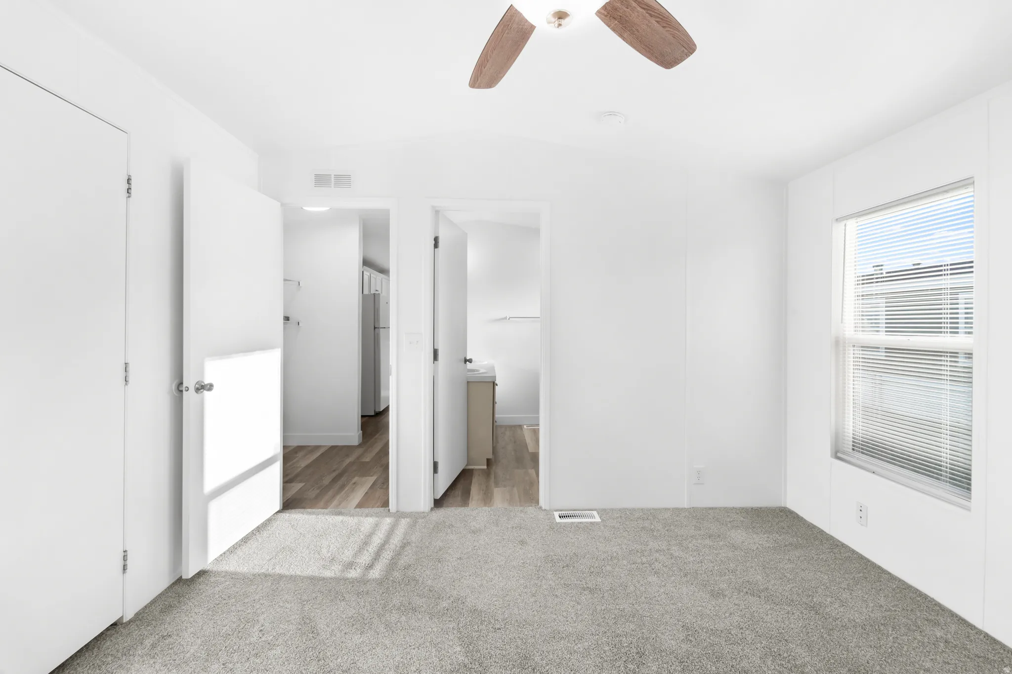 Neutral white interior featuring light gray carpeting, a window with horizontal blinds, and a ceiling fan with wood-finish blades