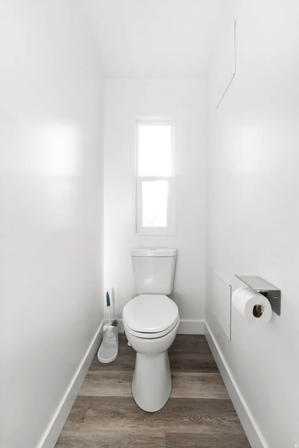 Windowed water closet featuring a two-piece toilet, wood-finish flooring, white baseboards, and a wall-mounted toilet paper holder